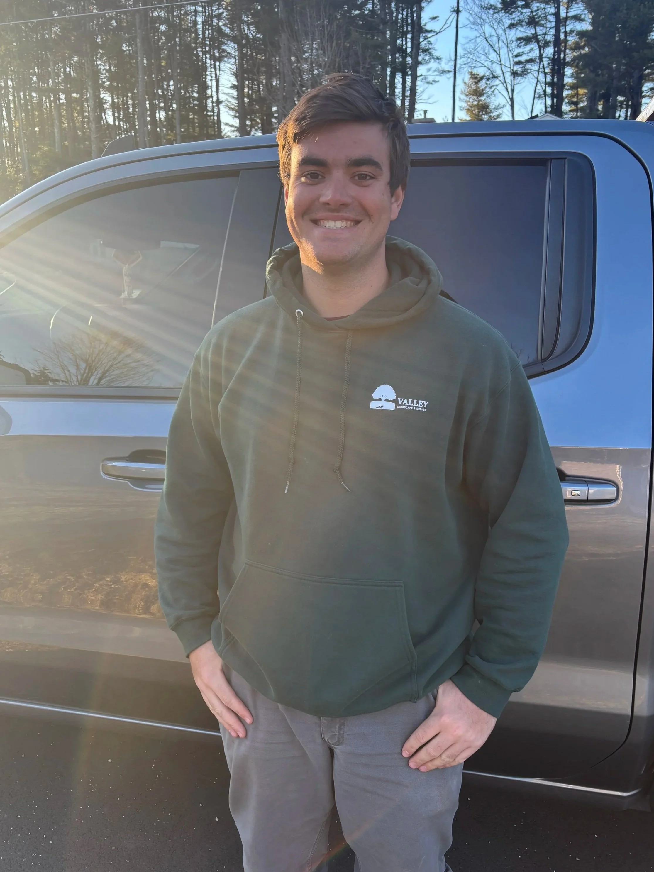 Young man standing next to a gray vehicle outdoors, smiling, wearing a dark green hoodie with a logo that says "Valley Landscapes & Design," with sunlight and trees in the background.