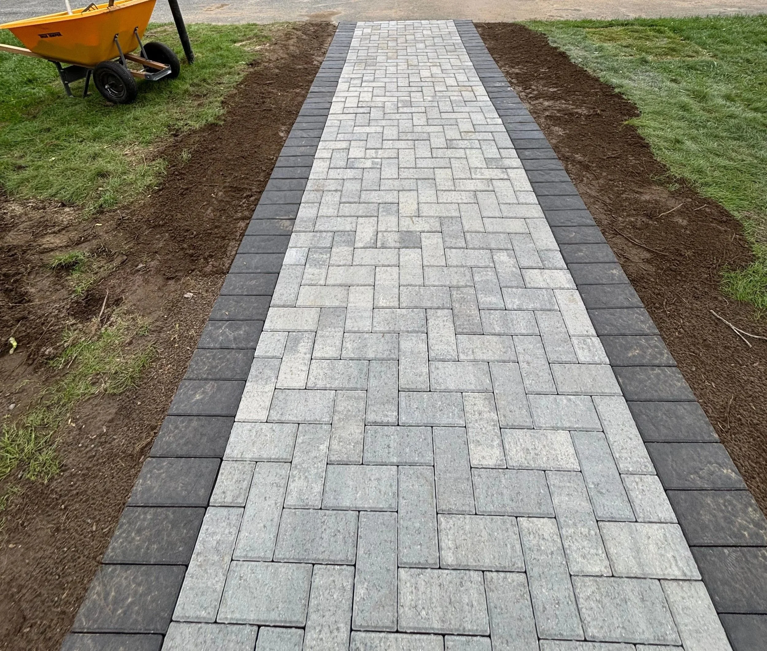 A newly paved brick walkway with a herringbone pattern, bordered by dark gray bricks, in process of installation on grass and soil.
