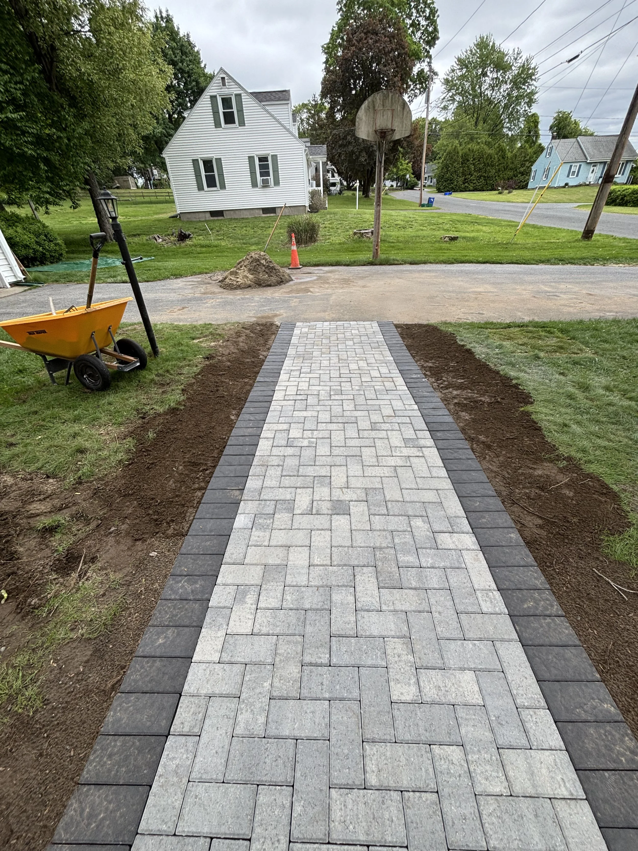 A newly paved pathway with light gray and dark gray bricks, bordered by dark gray bricks, leading to a residential street with houses, trees, and a basketball hoop in the background, surrounded by grass and construction materials.