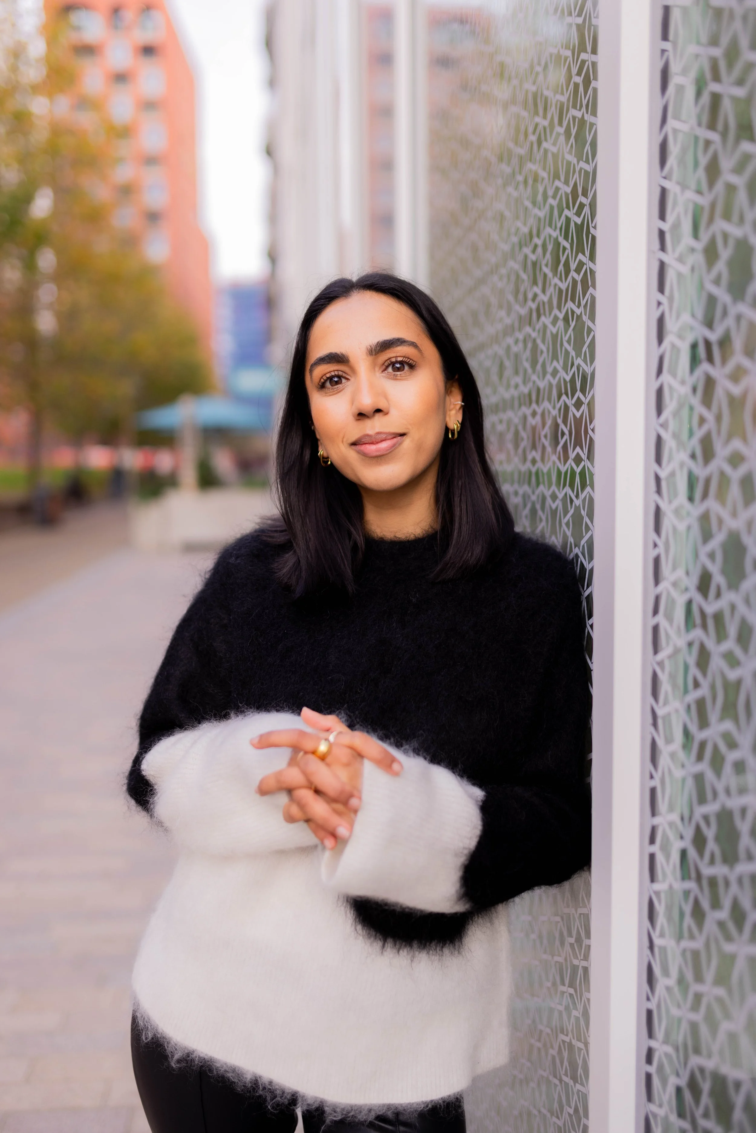 Woman with black hair and gold hoop earrings standing outdoors against a decorative wall, wearing a black and white fuzzy sweater.