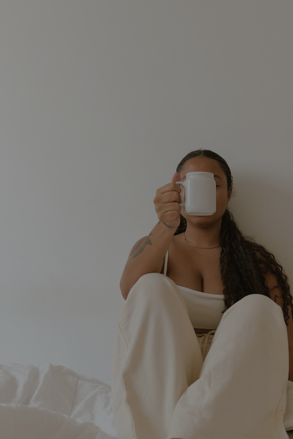 Woman with dark curly hair and tattoos sitting against a plain white wall, holding a white mug in front of her face.