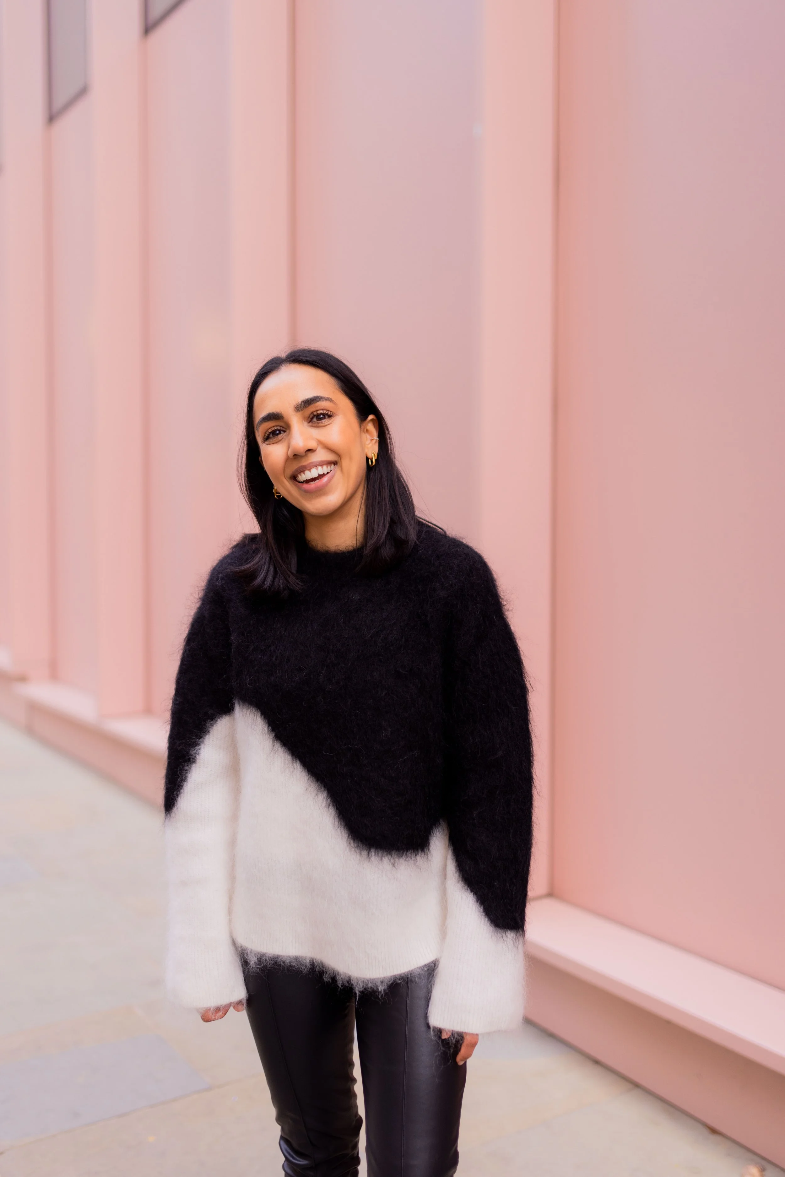 A woman with shoulder-length dark hair, wearing a black and white fuzzy sweater and black leather pants, smiling while standing in front of a pink wall.
