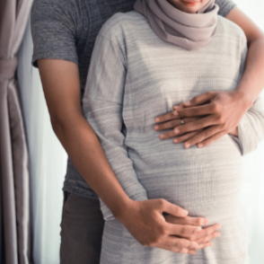 A pregnant woman with hands on her belly being embraced from behind by a partner.