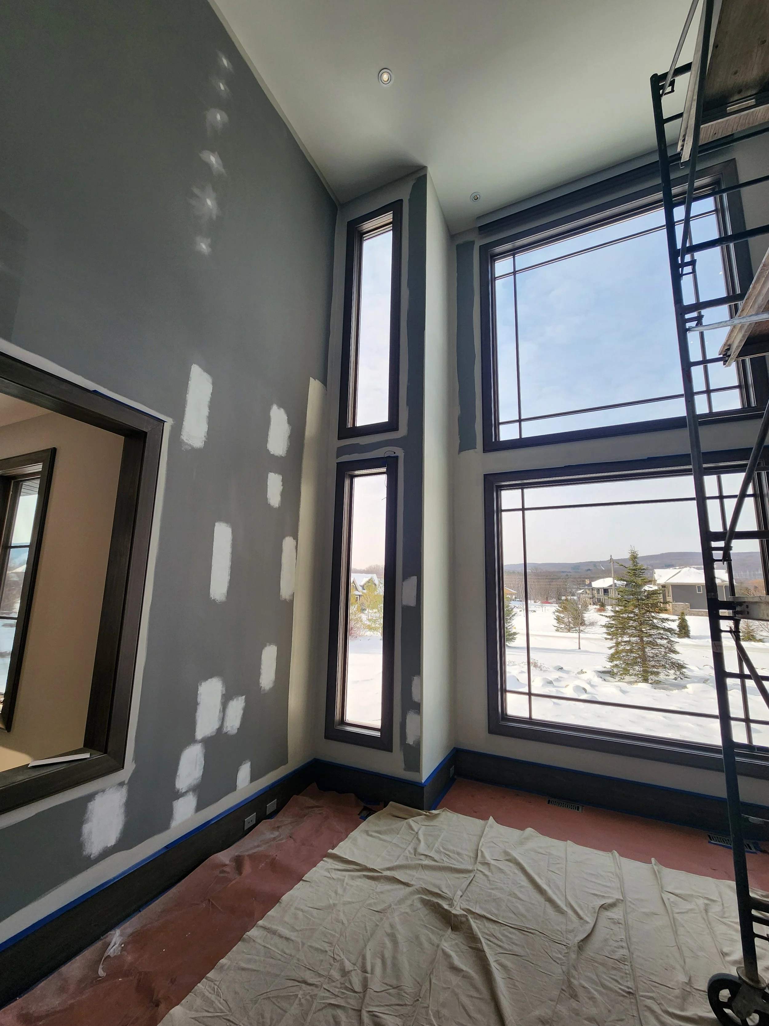 Interior of a room under construction with unfinished walls, large windows showing a snowy landscape outside, and construction equipment.