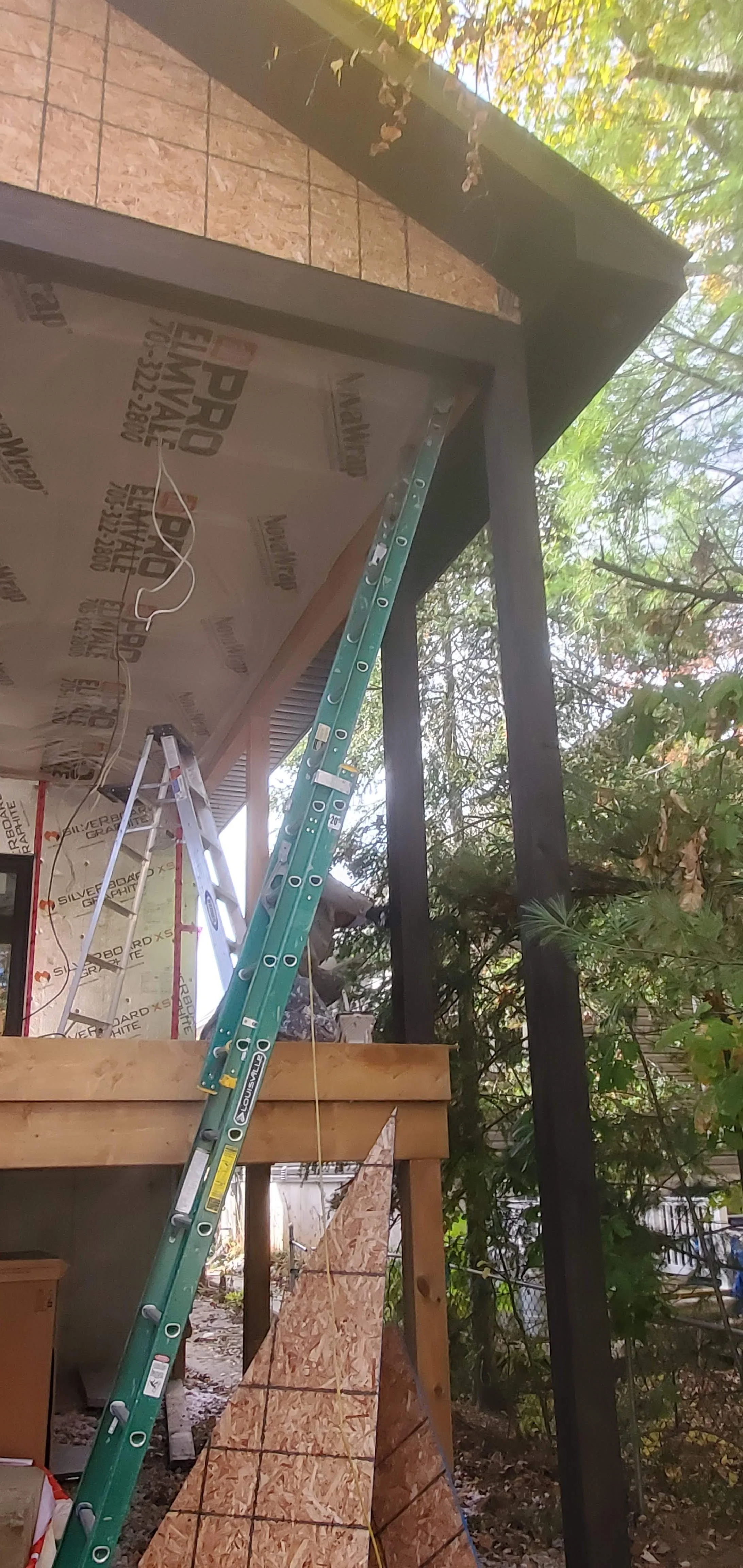 Construction or renovation in progress with a ladder, wood panel, and building materials under an overhang, surrounded by trees.