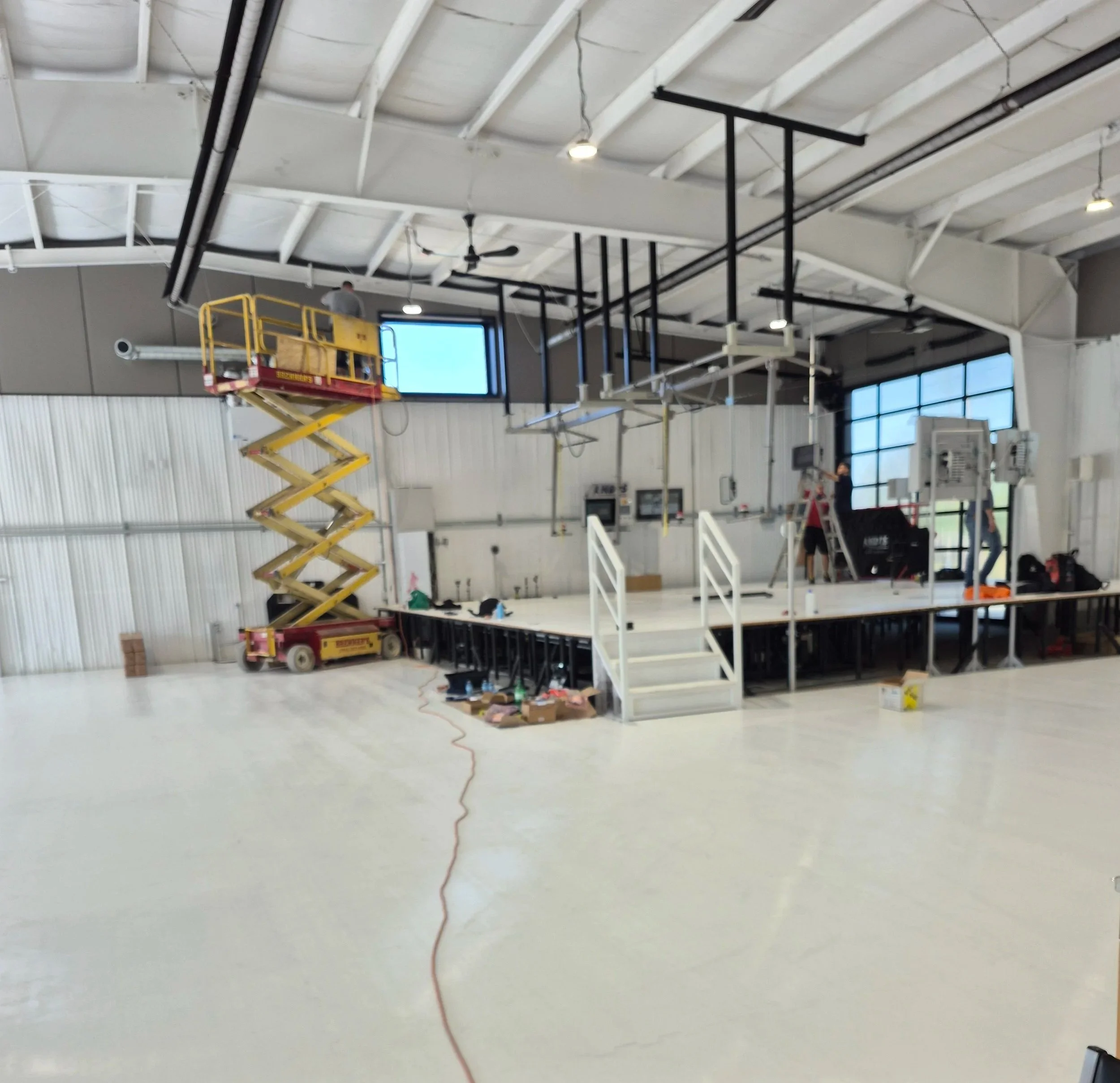 Inside a large industrial space with a stage under construction, featuring a yellow scissor lift, tools, and workers setting up equipment.