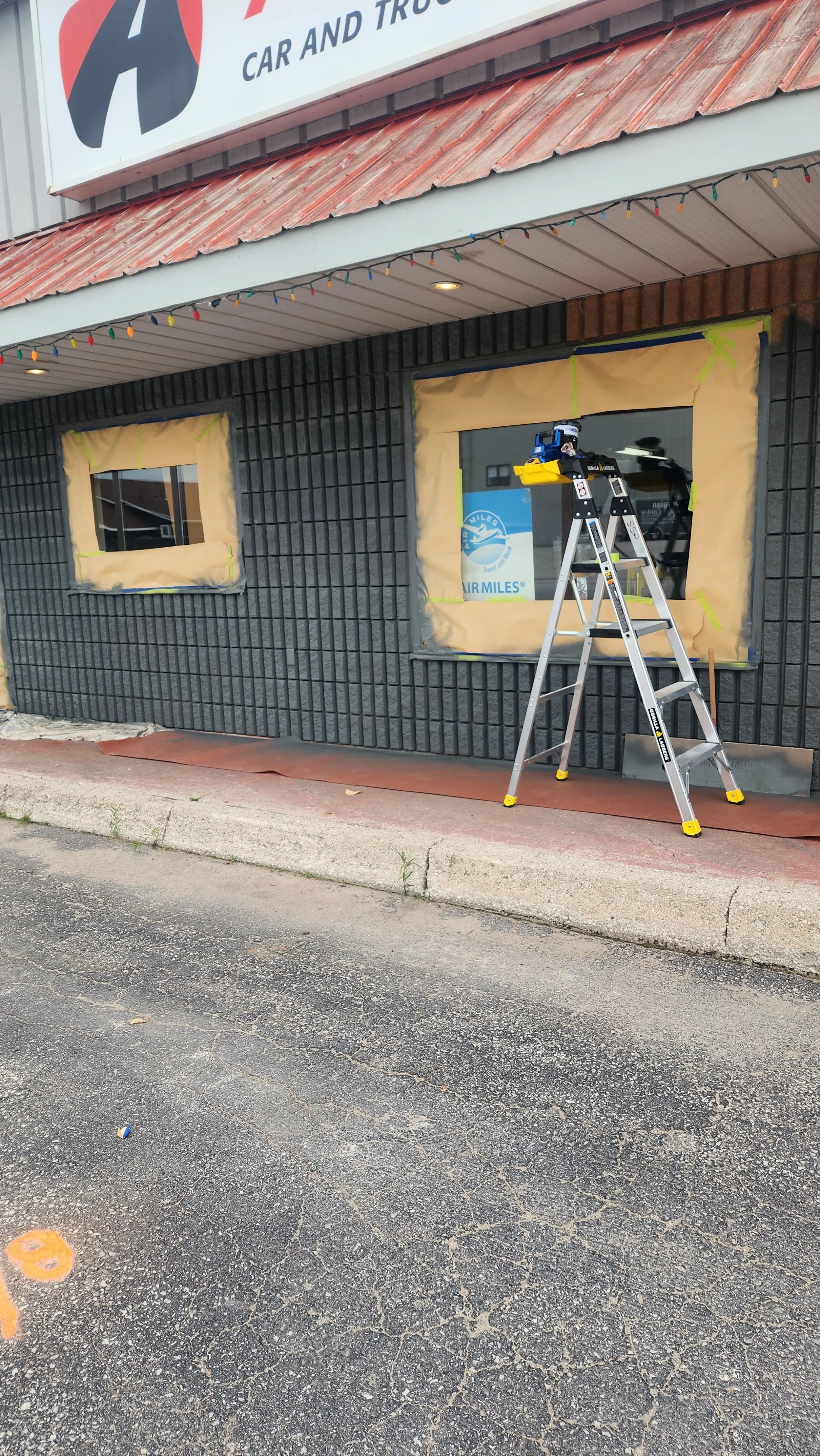 Exterior of a business under construction or renovation, with black brick wall, two windows covered with beige painter's paper, and a folding ladder with a power tool on top, outside a store selling cars and trucks.