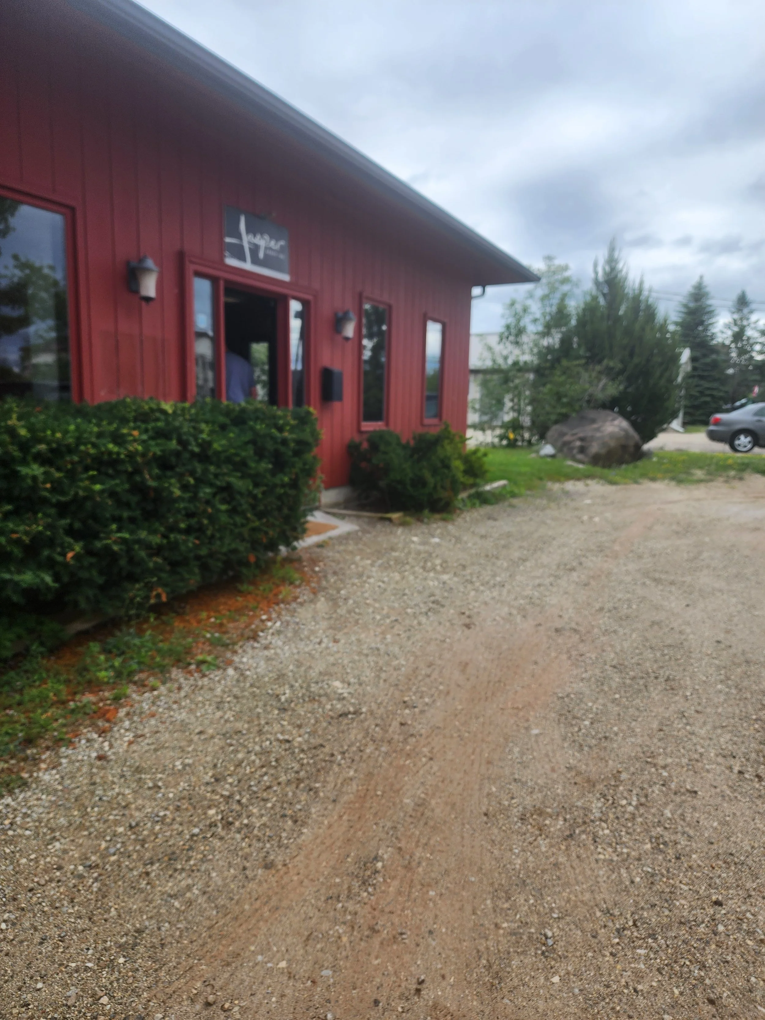 Red building with a sign above the door, surrounded by bushes, with a gravel driveway and a few cars parked nearby, under a cloudy sky.