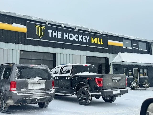 Snow-covered parking lot in front of a building with a sign that reads 'The Hockey Mill' with parked cars, including an SUV and a pickup truck.