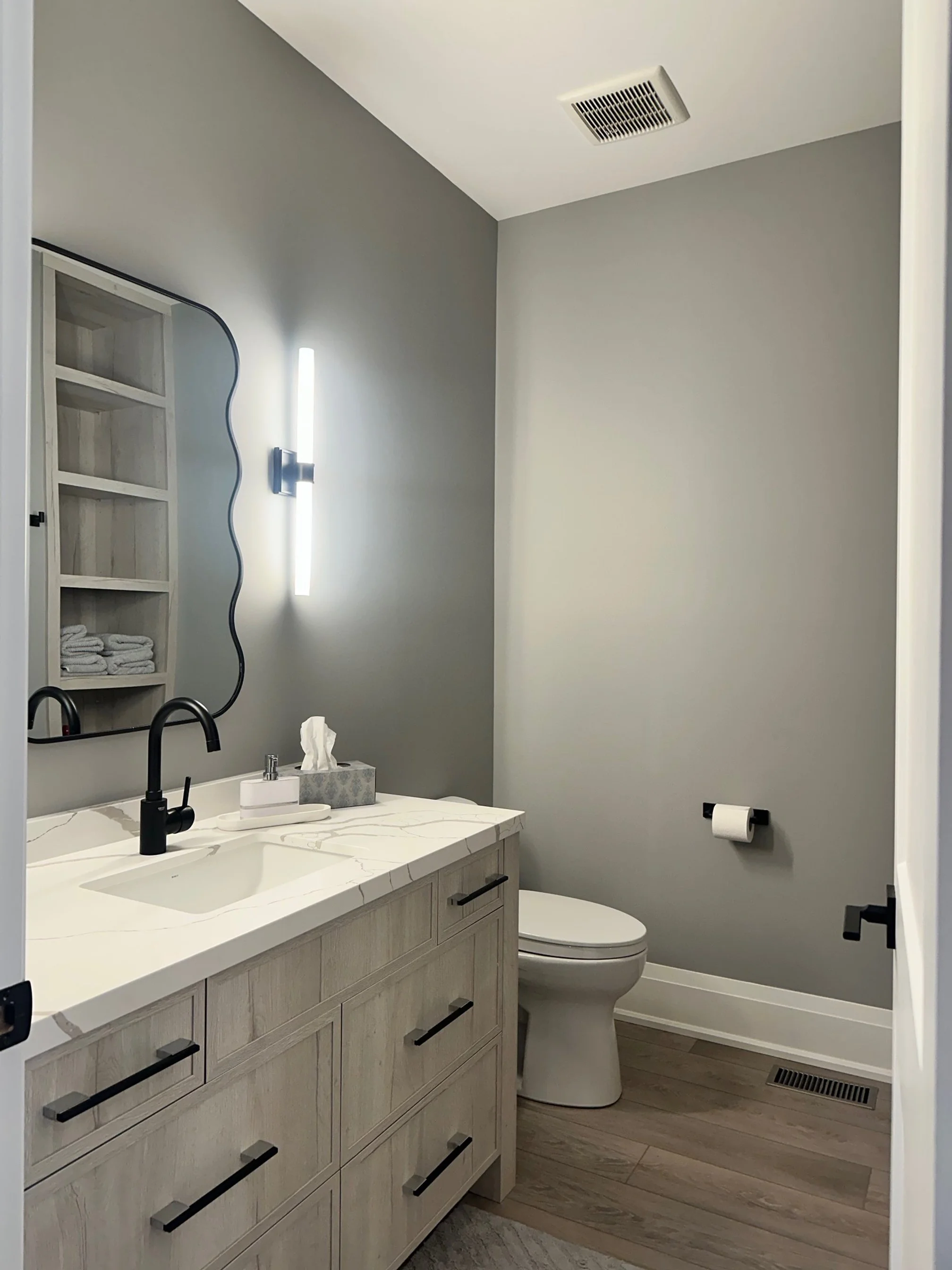 Modern bathroom with a white marble vanity, black faucet, gray walls, a mirror, sconces, a toilet, toilet paper holder, wooden flooring, and a vent on the ceiling.