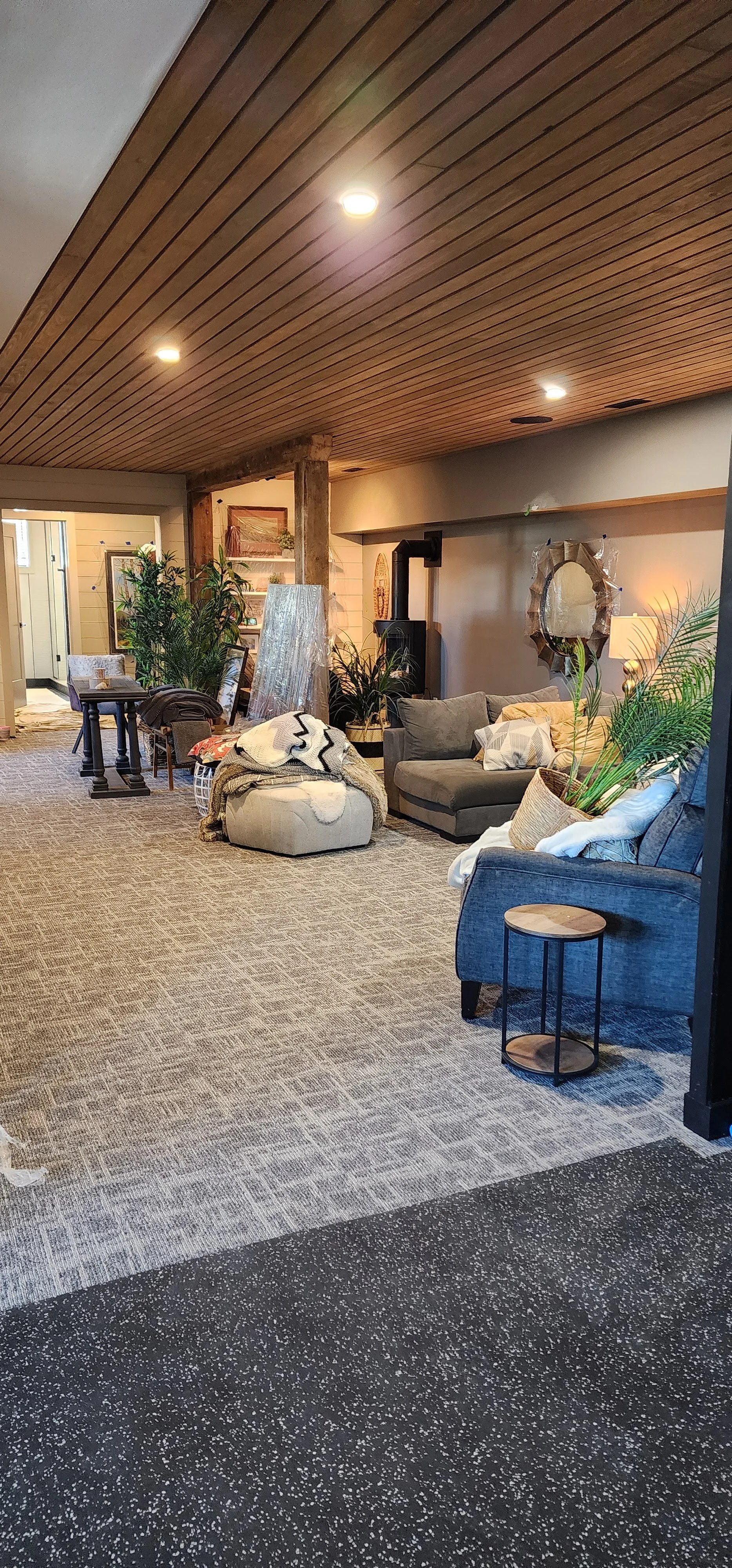 A cozy living room with wooden ceiling, grey and beige sofas, a round pouf with a blanket, potted plants, a small side table, and wall decor, illuminated by ceiling lights.