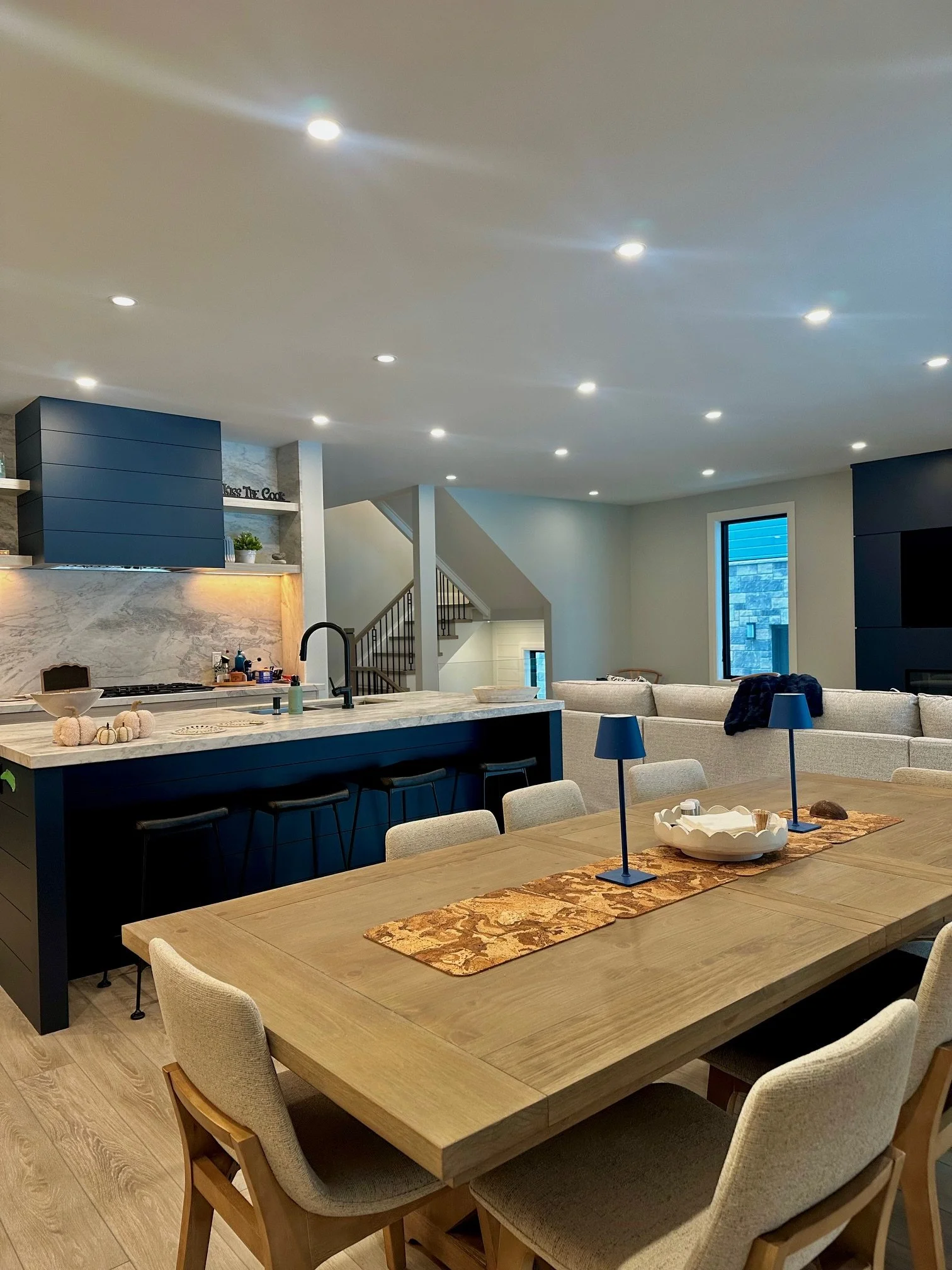 A spacious modern living room and kitchen area with hardwood floors, a large dining table with six upholstered chairs, a kitchen island with bar stools, a blue and white color scheme, and recessed ceiling lights.