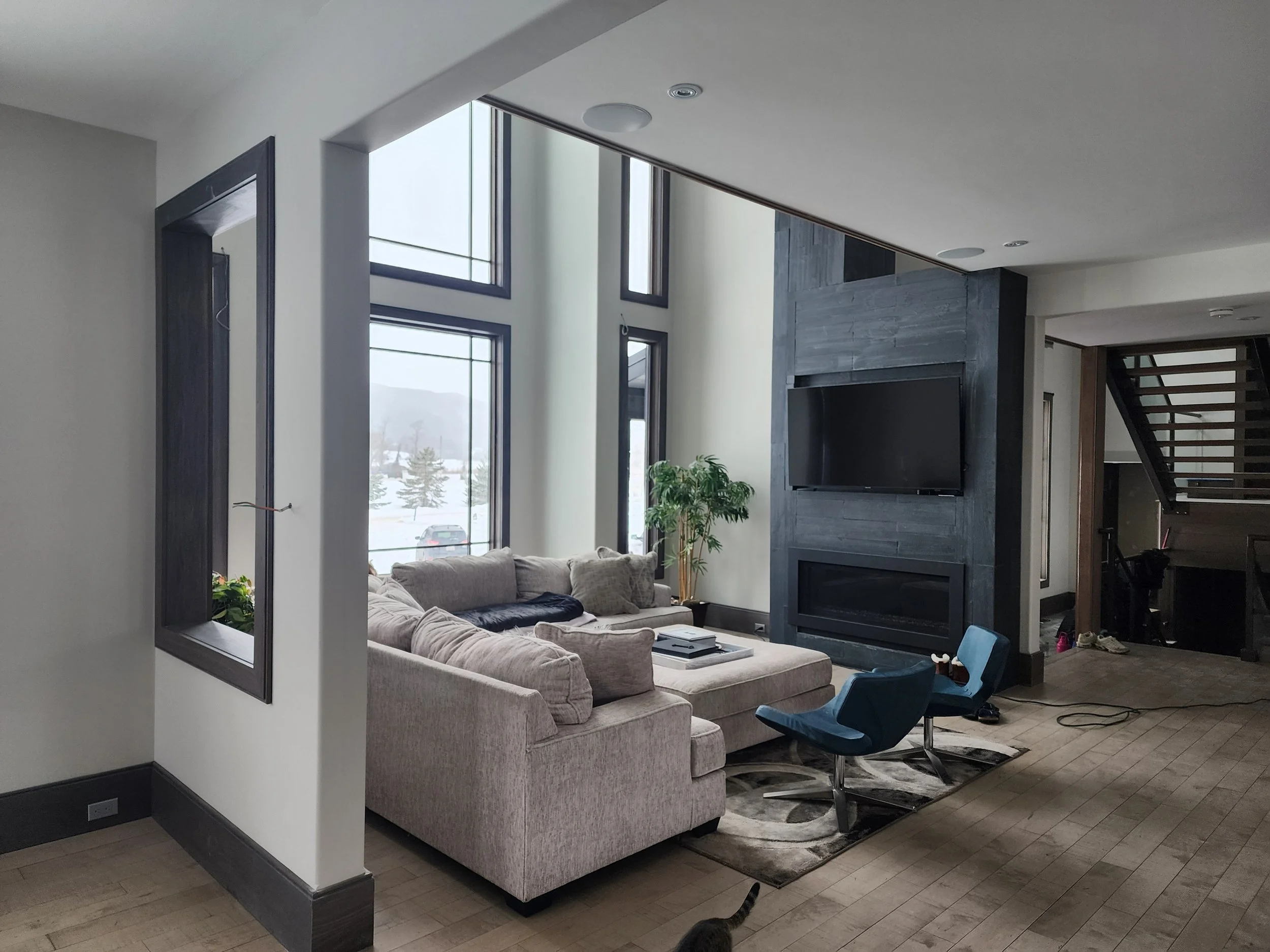 Living room with a beige sectional sofa, a blue chair, a large wall-mounted TV above a black fireplace, and large windows showing a snowy landscape outside.