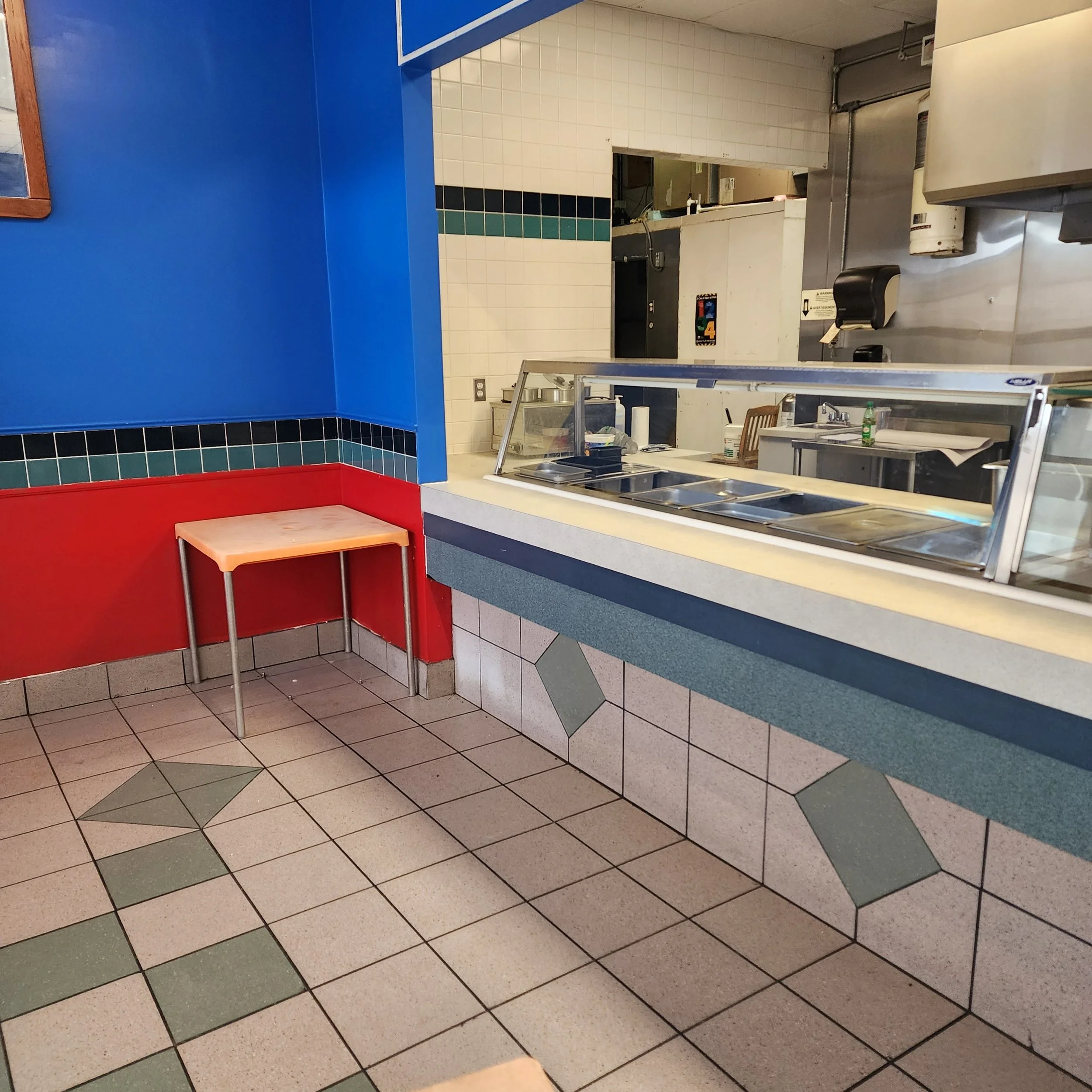 Empty fast-food restaurant counter with a colorful corner seating area, featuring blue and red walls, tiled floor, and a glass display counter for food service.