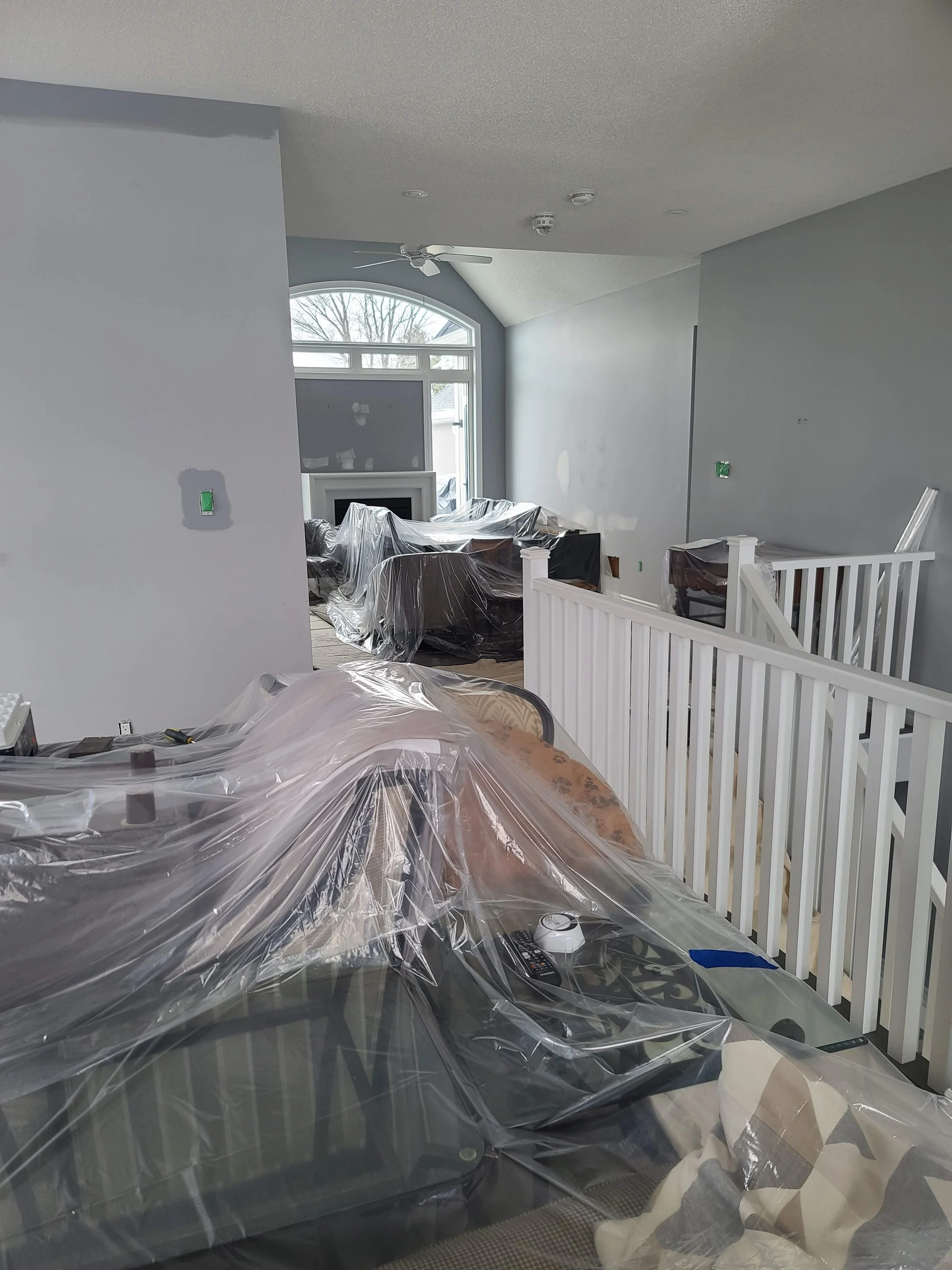 Living room under renovation with furniture covered in plastic, a ceiling fan, a fireplace, and a large window.