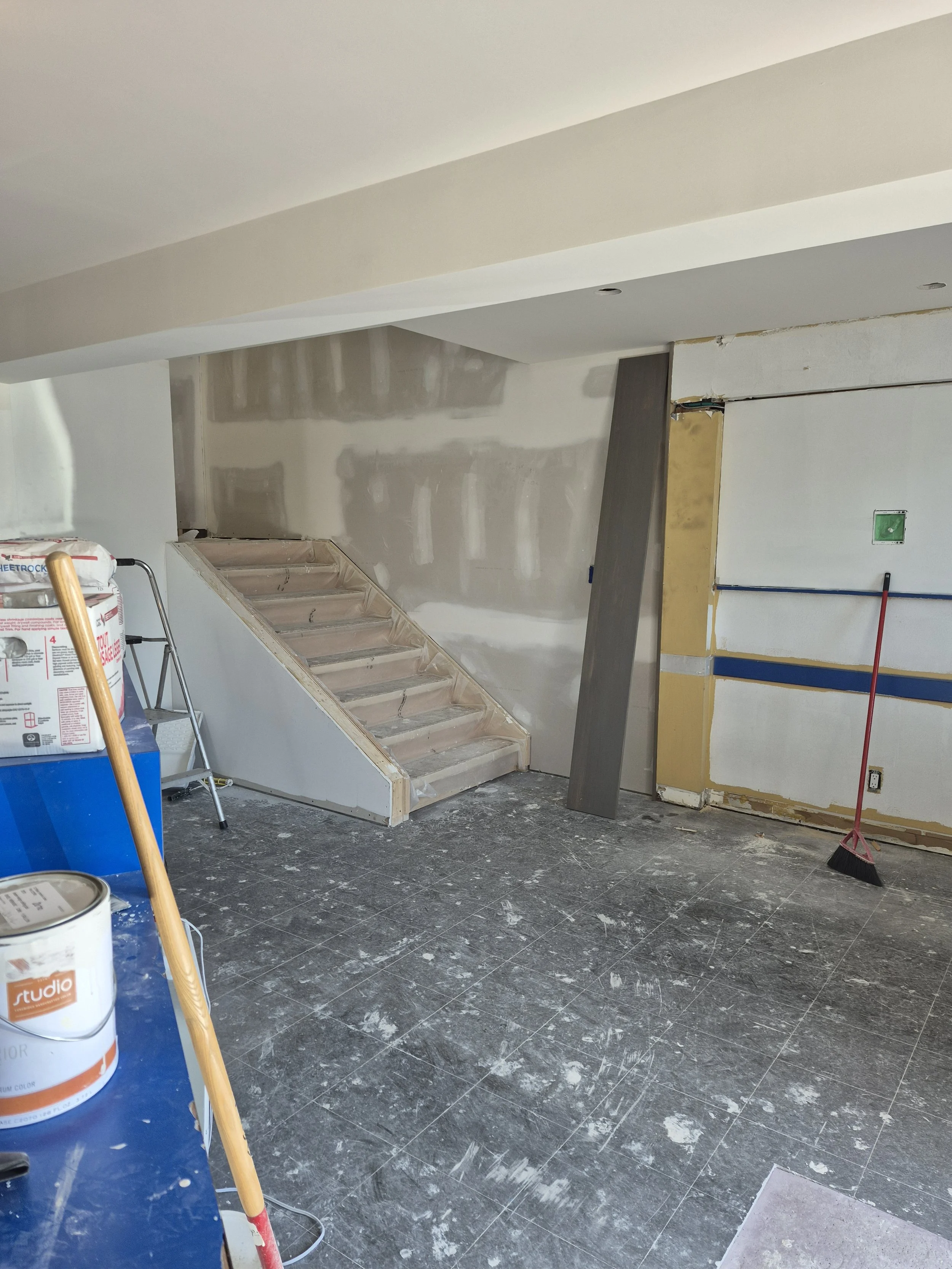 Interior of a construction site with partially finished walls, a staircase, and cleaning tools on a dusty black tile floor.
