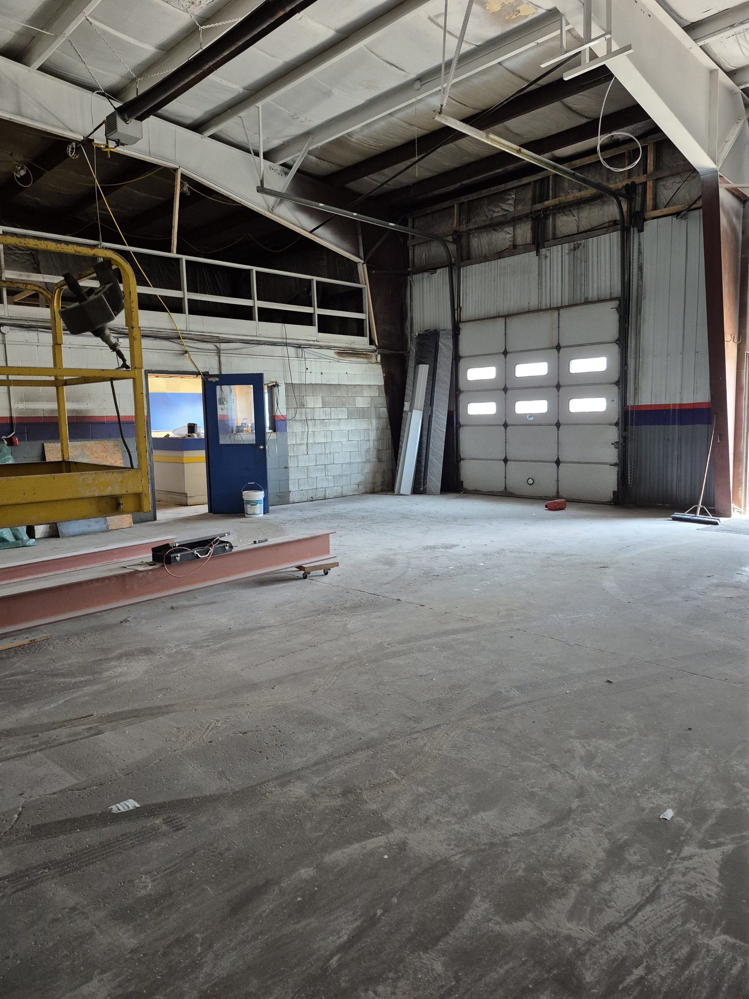 Empty industrial or warehouse space with a large garage door, some construction materials, and equipment scattered around.