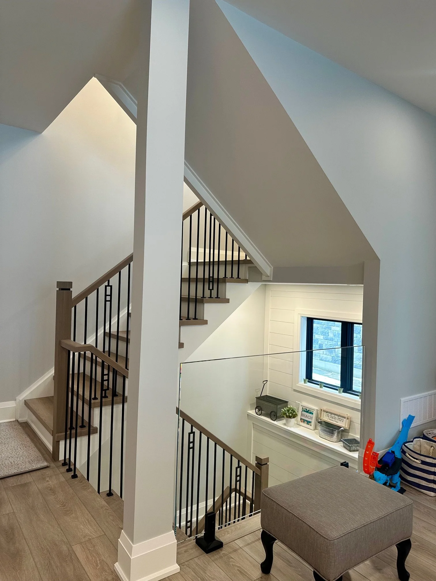 Interior view of a multi-level staircase with black railings, white walls, and a window overlooking an outdoor scene. A small beige ottoman is in the foreground, with toys and decor items on a ledge below the window.