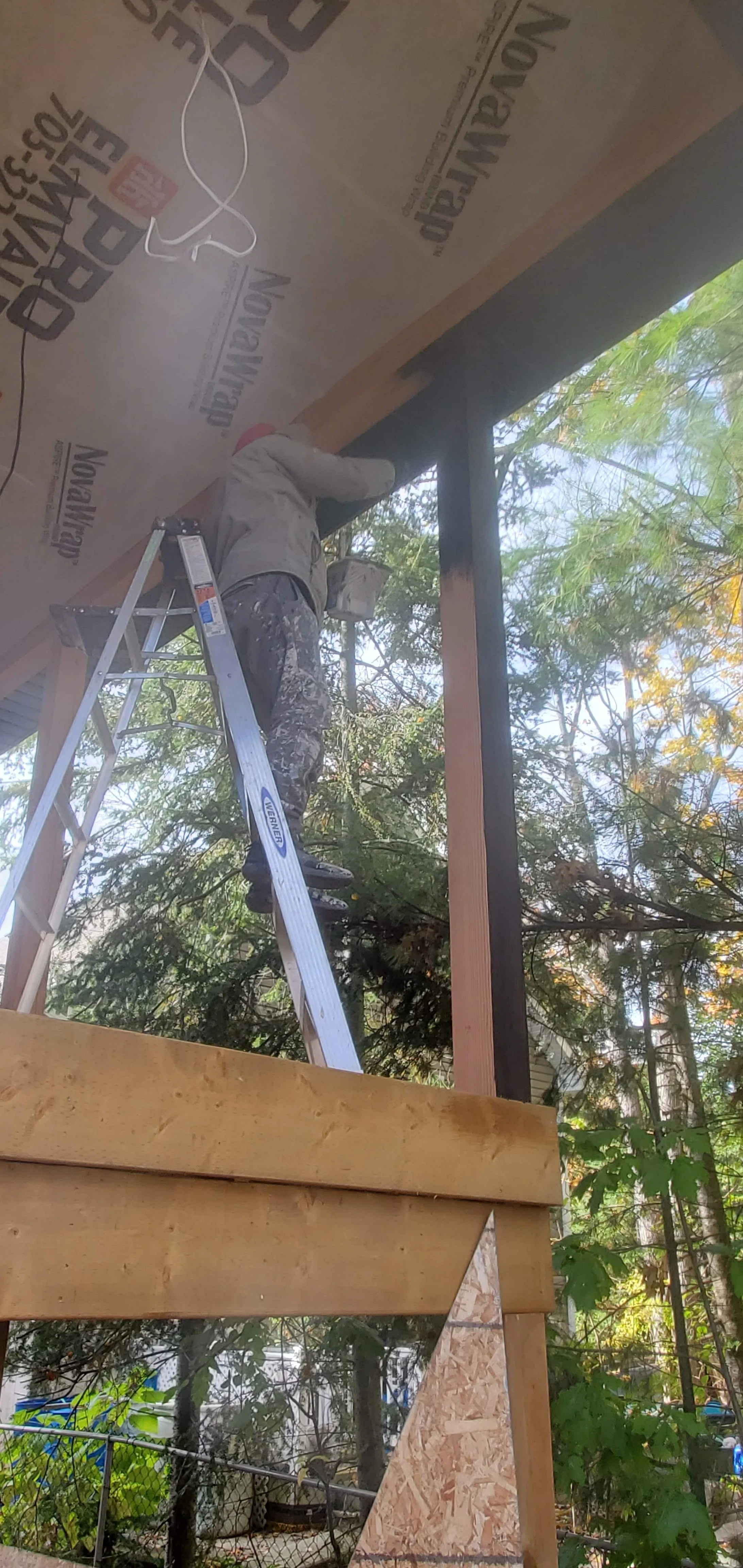 A person is standing on a ladder, working on the underside of a roof eave or soffit in a construction area surrounded by trees, with tools and construction materials nearby.