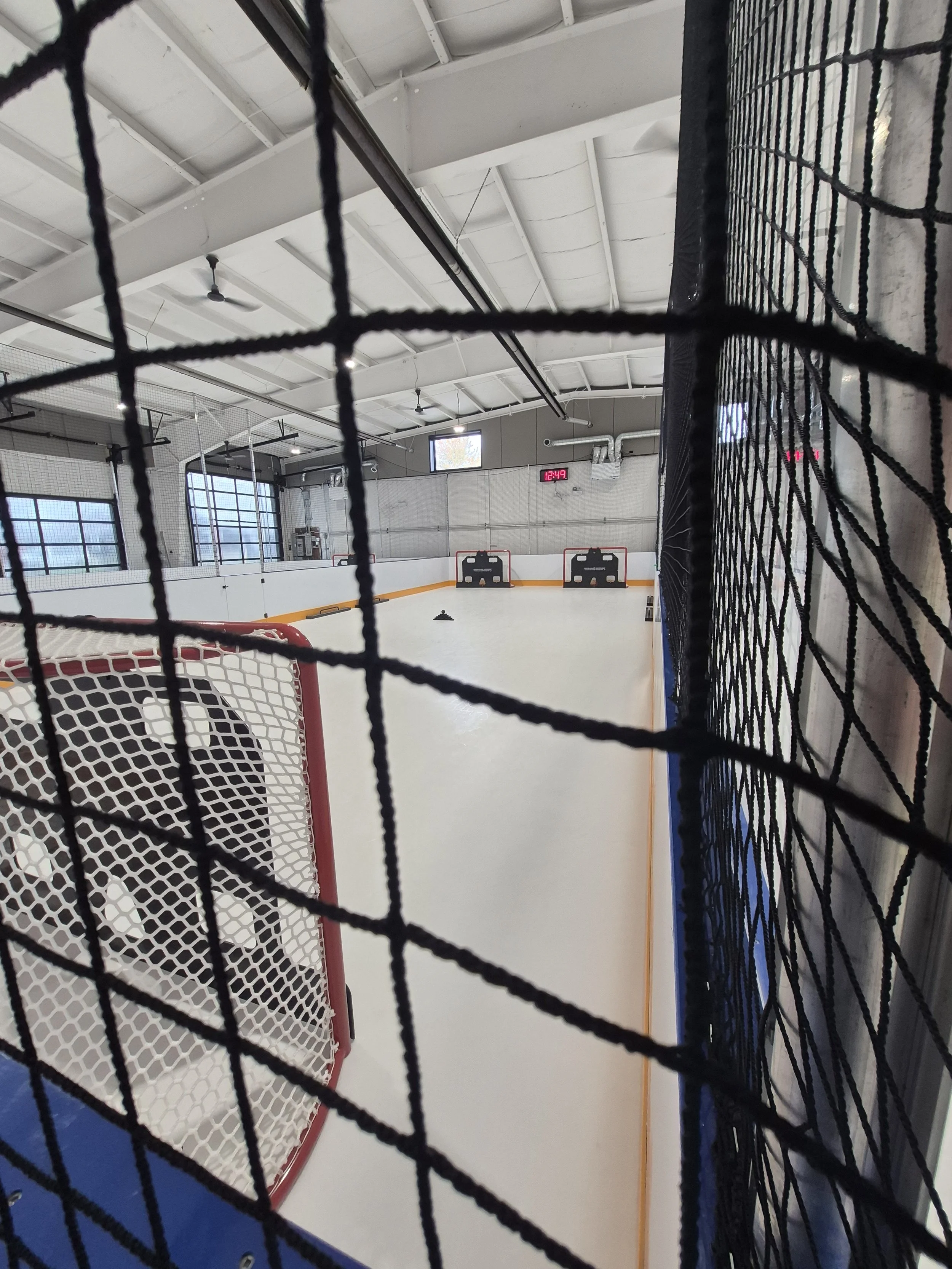 An ice hockey rink viewed through a black net. The rink is empty, with hockey goals at each end and a digital clock showing 12:49. Overhead lights and a ceiling fan are visible.