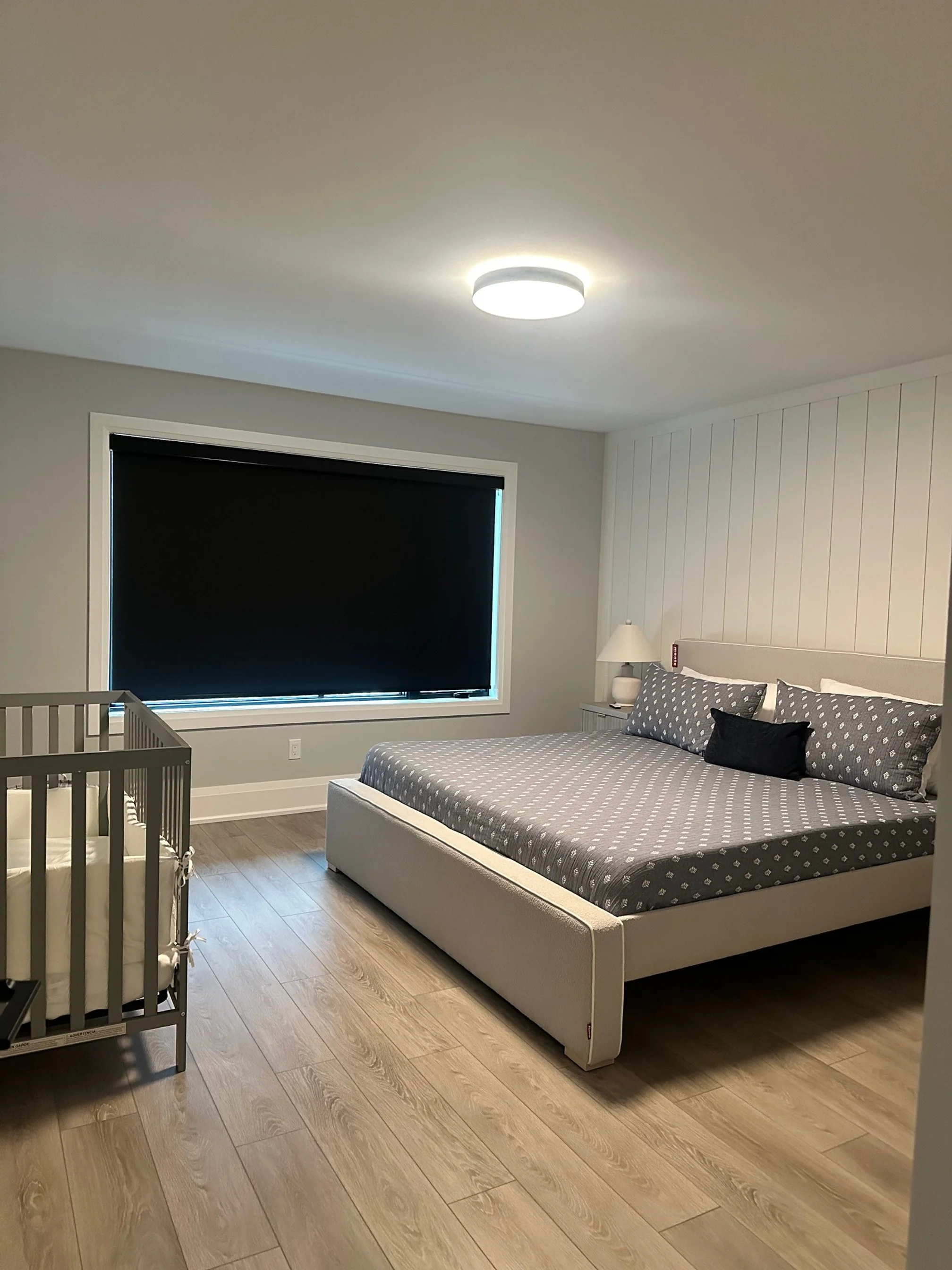 A bedroom with a large bed featuring a patterned gray bedspread and pillows, a nightstand with a lamp, a window with a closed black blind, and a baby crib on the left side, all with light-colored walls and wooden flooring.