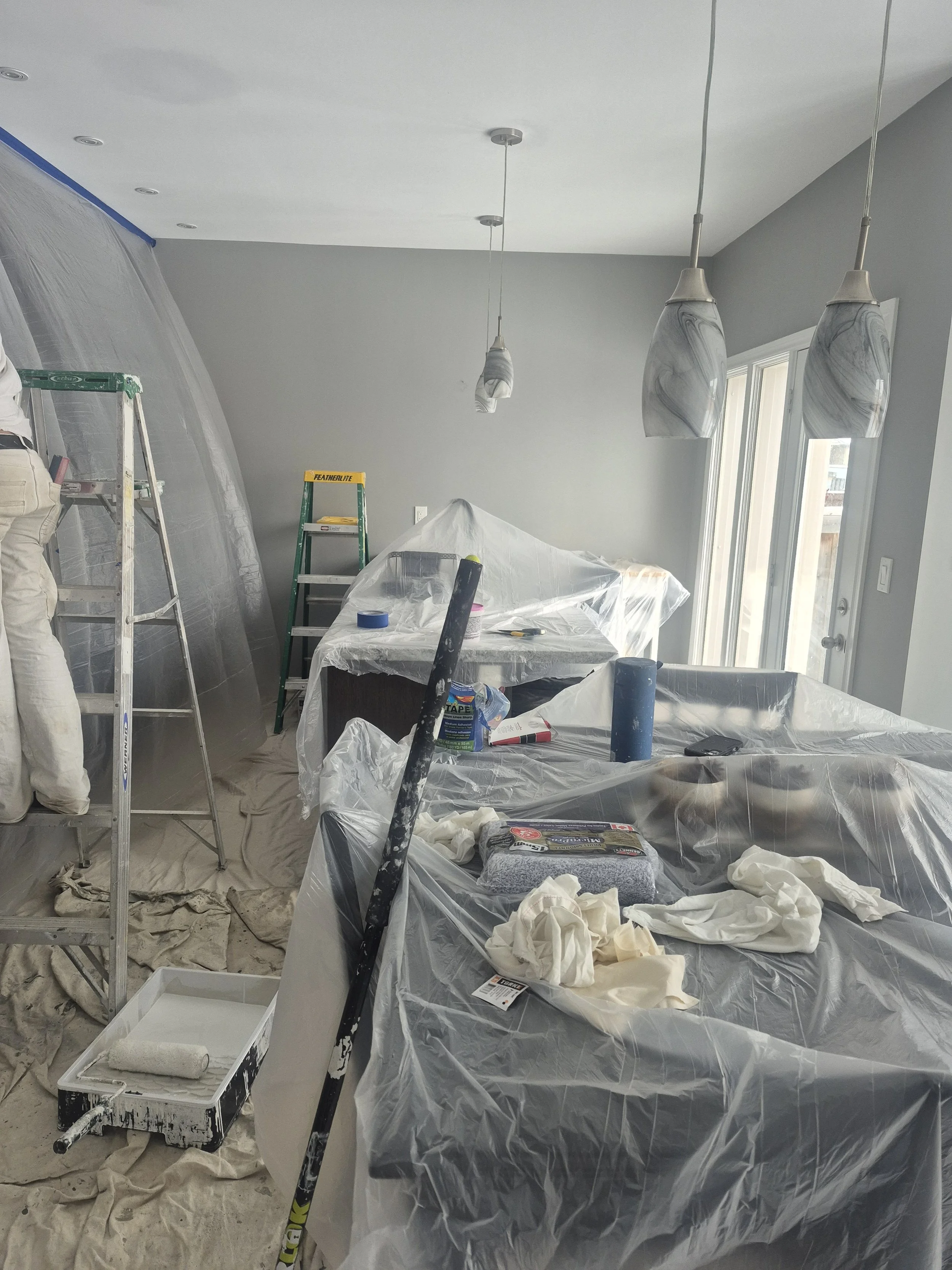 A room under painting renovation with plastic sheeting covering furniture and walls, ladders, paint cans, brushes, and painting supplies.