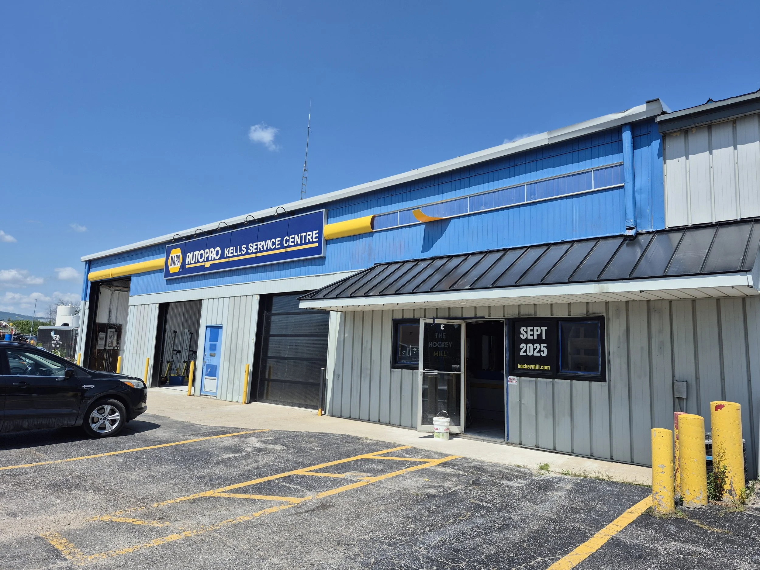 Exterior of a building with a sign that reads 'NAPA Autopro Kells Service Centre.' The building has a blue and gray facade, with a large yellow and gray pipe decoration above the sign. There are a few cars parked in front, and the parking lot has yel