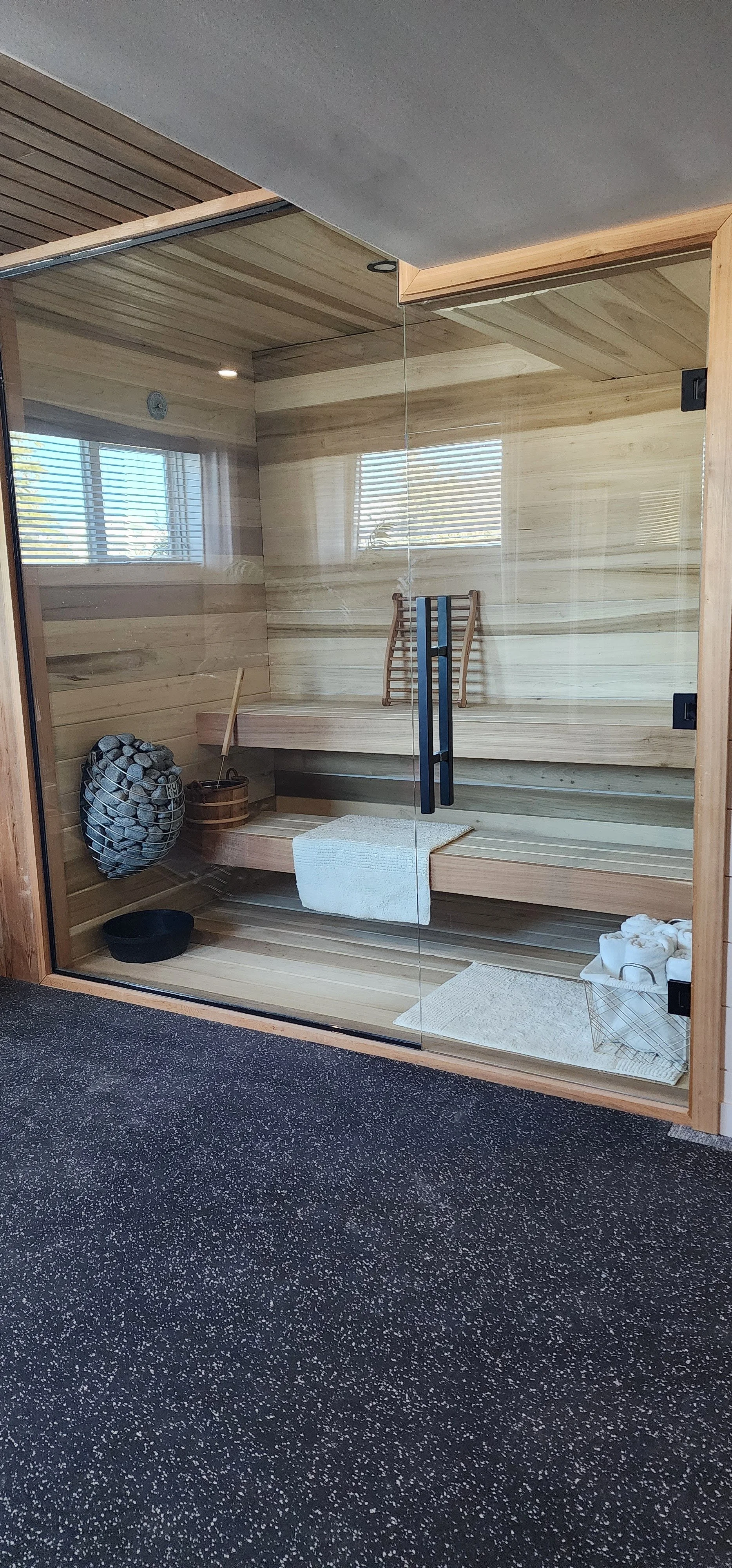 A modern wooden sauna with glass door, wooden benches, sauna accessories like a bucket, ladle, and towels, illuminated by natural light through windows.
