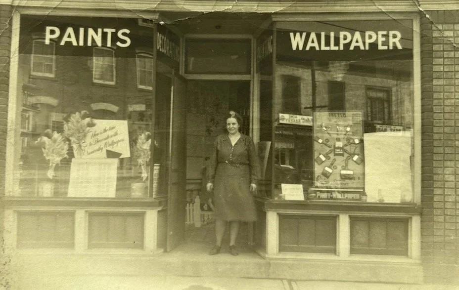A vintage sepia photo of a woman standing in the entrance of a store with large windows displaying paintings and wallpaper. The store has signs that read 'PAINTS' and 'WALLPAPER' in bold letters.