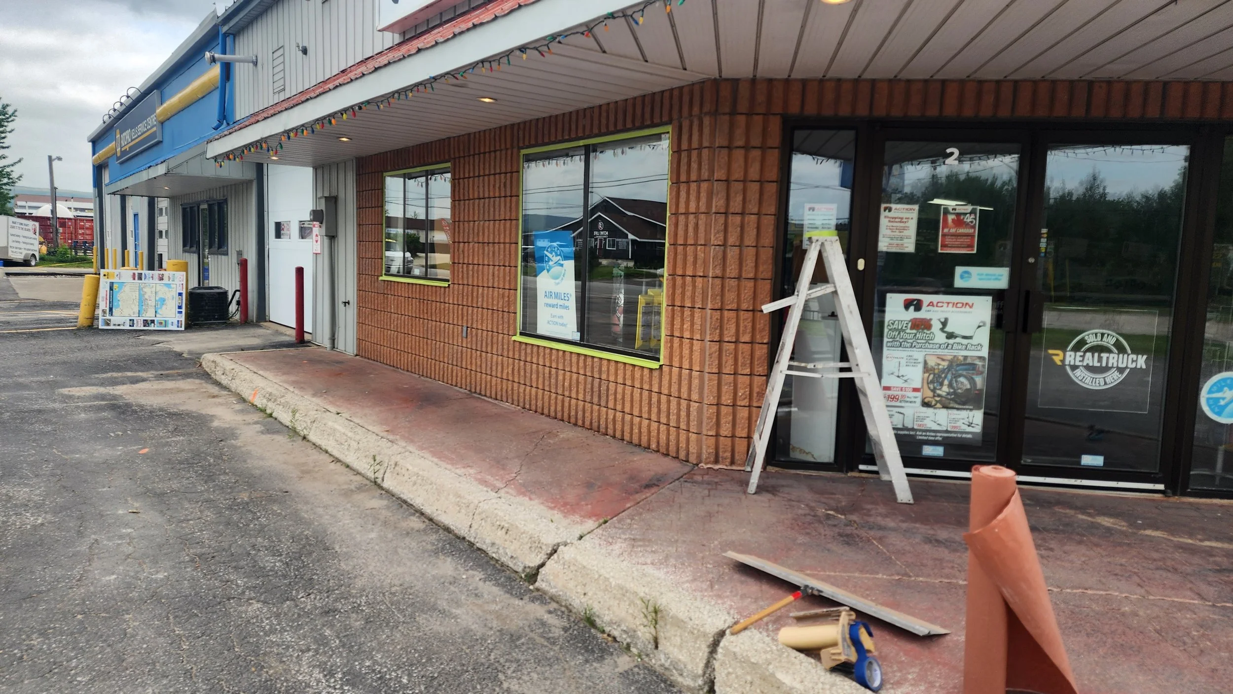 A storefront with a brick exterior and large windows, one with a sign for RealTruck. There is a ladder and construction materials outside, indicating ongoing work. The parking lot is partially visible, with a few notices on the door and windows.