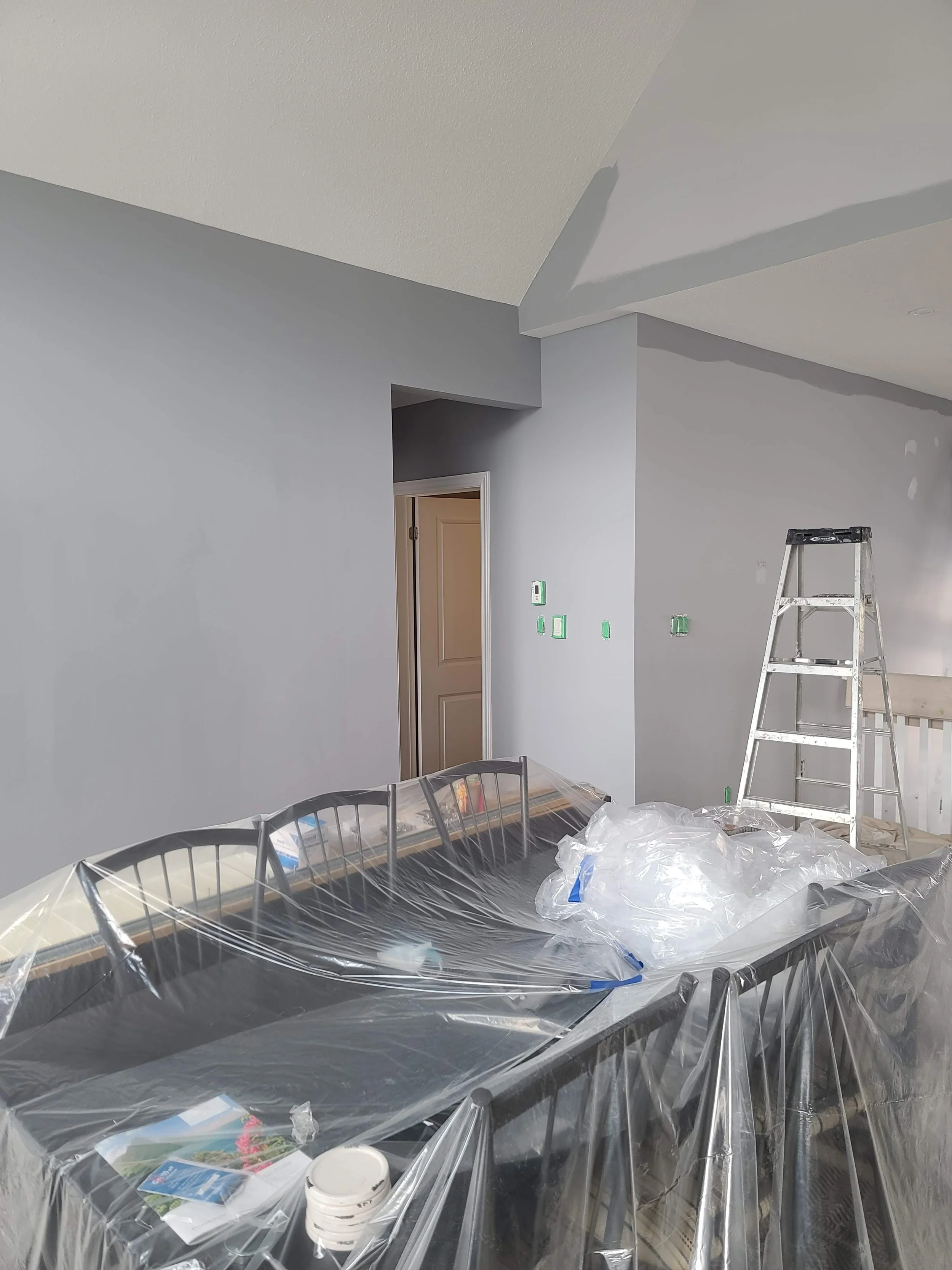 Interior of a room under renovation with light gray walls, a silver step ladder, a table covered in plastic, and a doorway in the background.