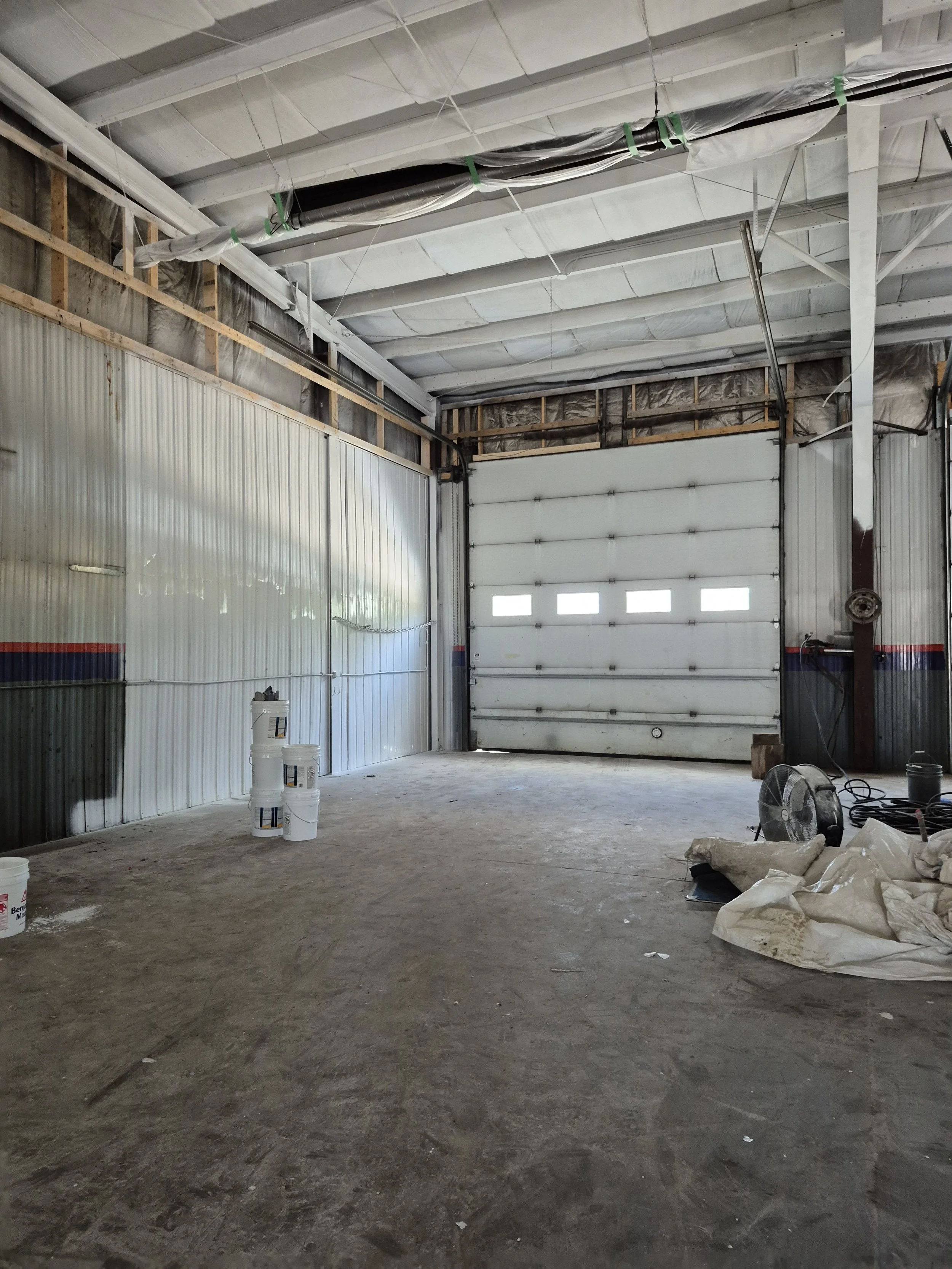 Interior view of an unfinished garage or warehouse with concrete floor, metal walls, and a large garage door. Construction materials and tools are scattered on the floor.