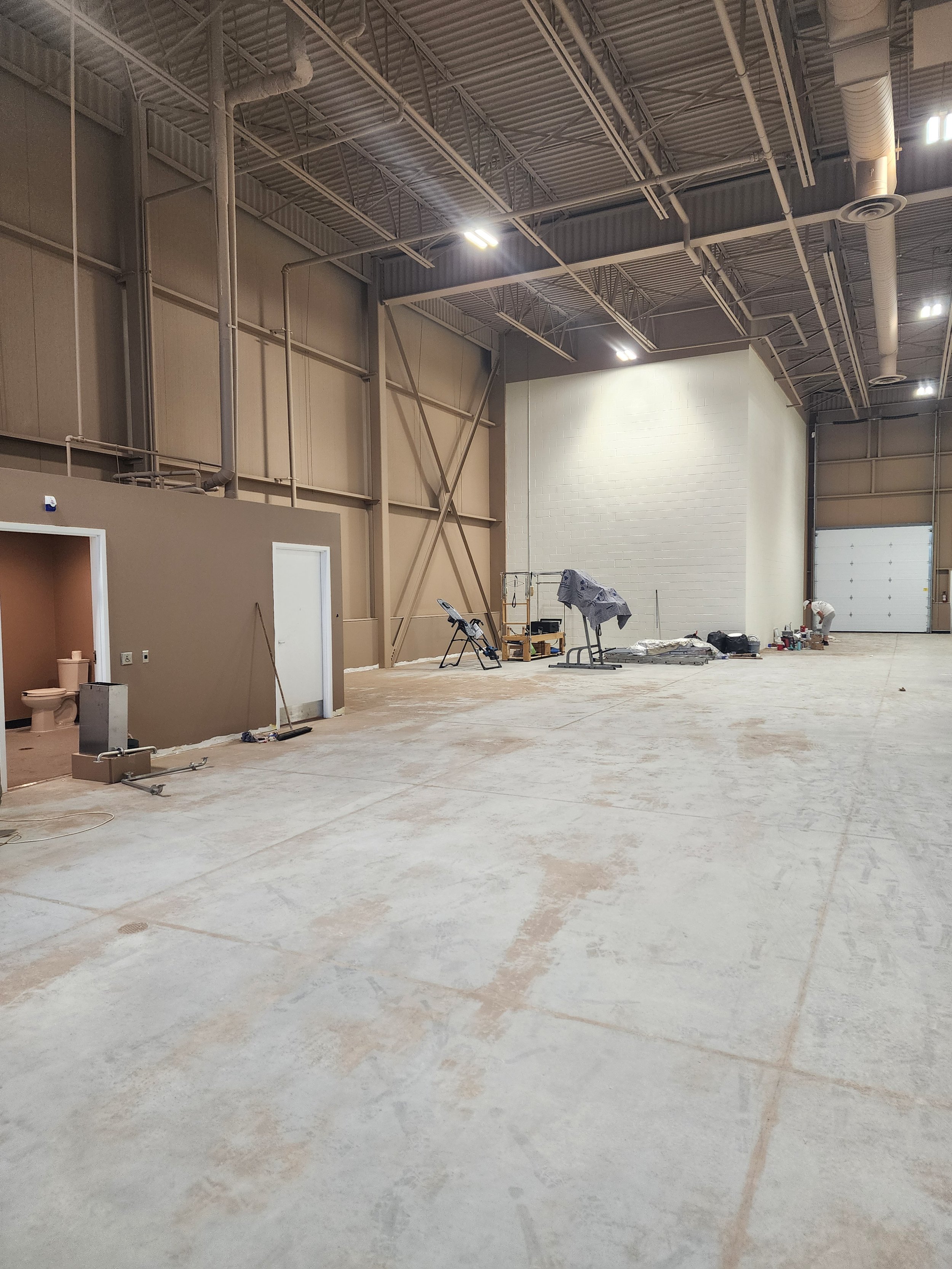 Inside a large industrial warehouse under construction with unfinished concrete floors, exposed metal ceiling beams, and some construction tools and materials scattered around. A small room with a toilet is visible on the left, and a worker is workin