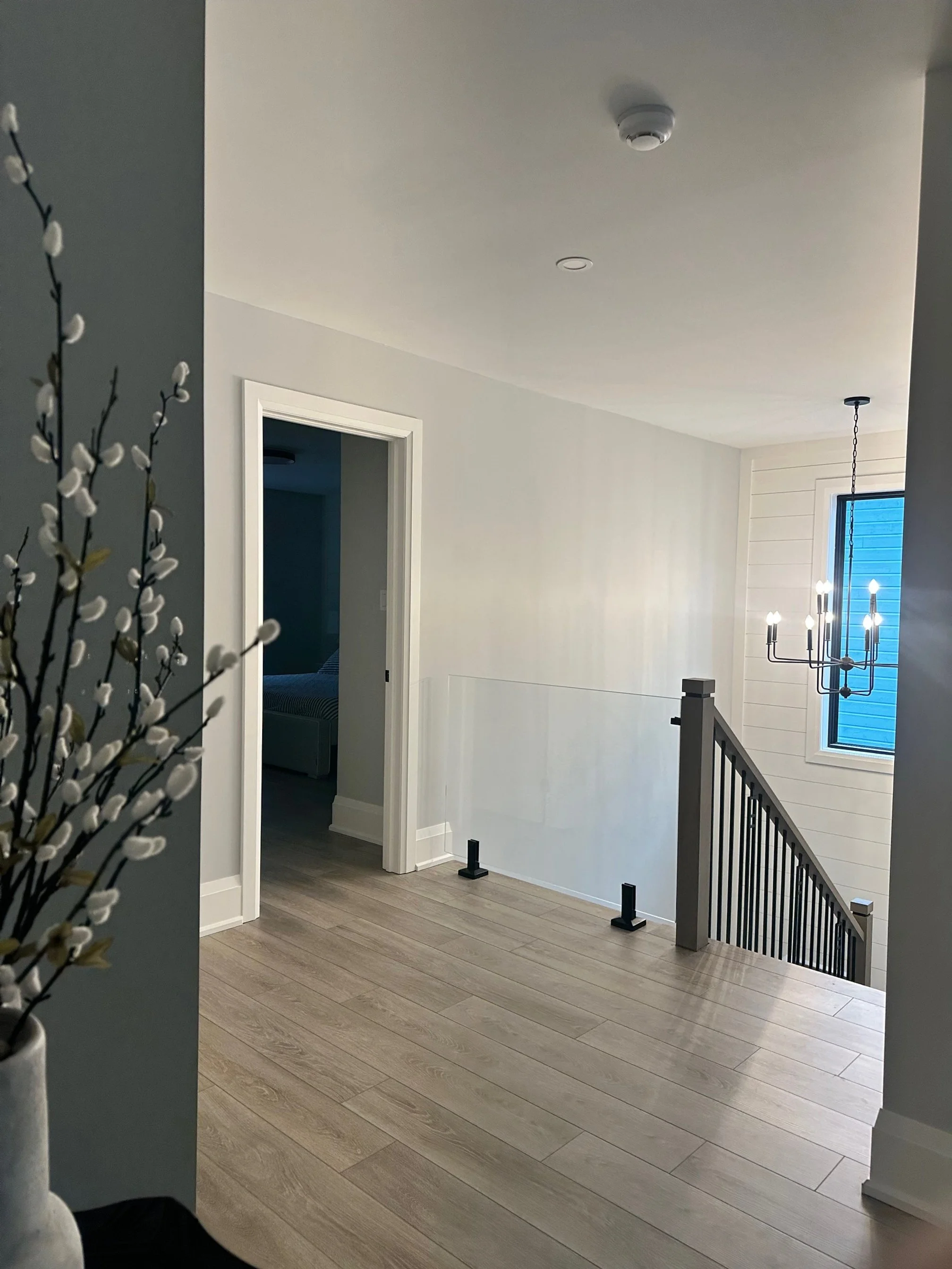 Interior view of a stairway landing with a chandelier, window, and door leading to a bedroom, with light-colored wooden floors and white walls.