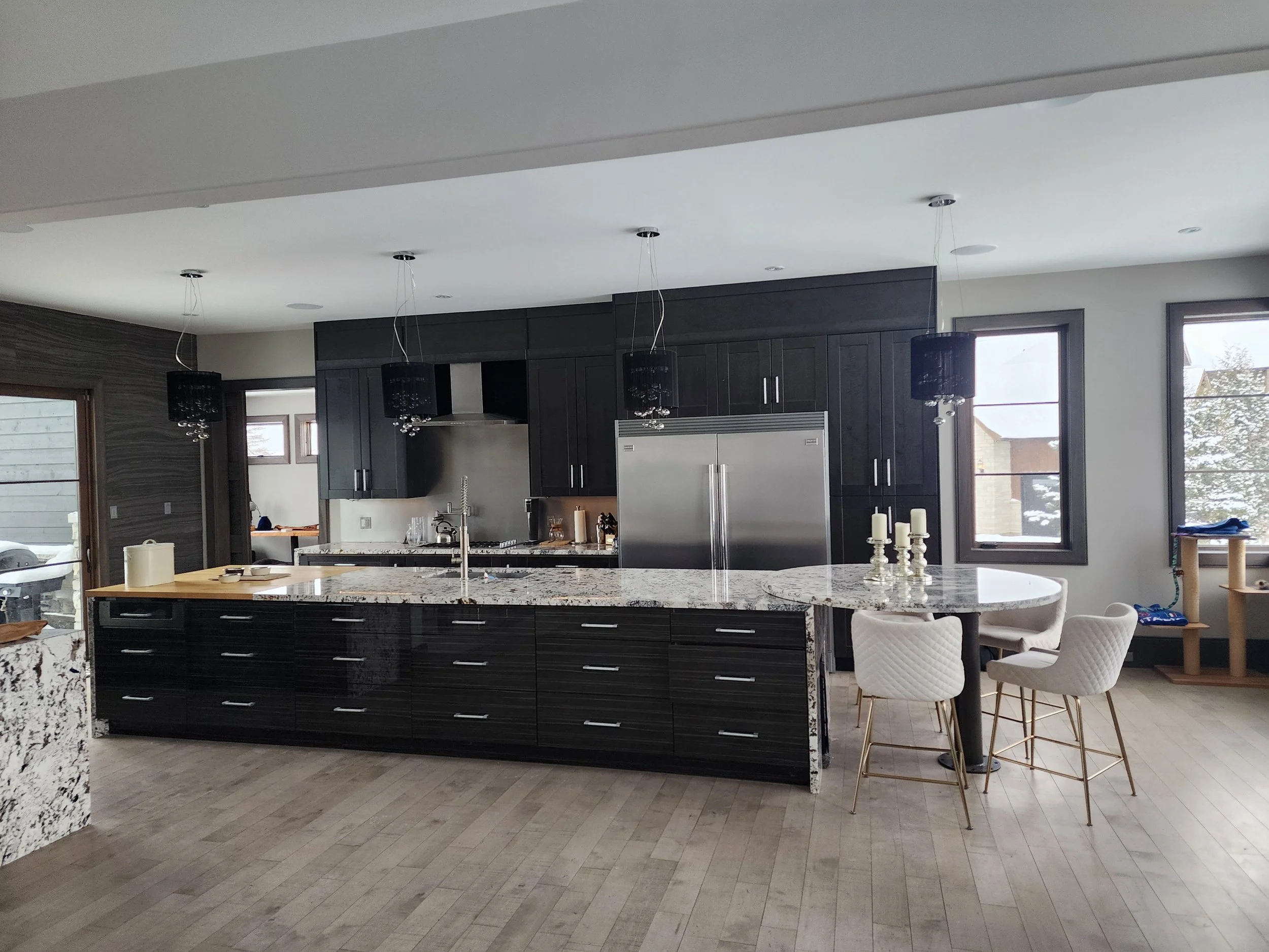 Modern kitchen with black cabinetry, granite island, and dining area with four white chairs, large windows, and pendant lighting.