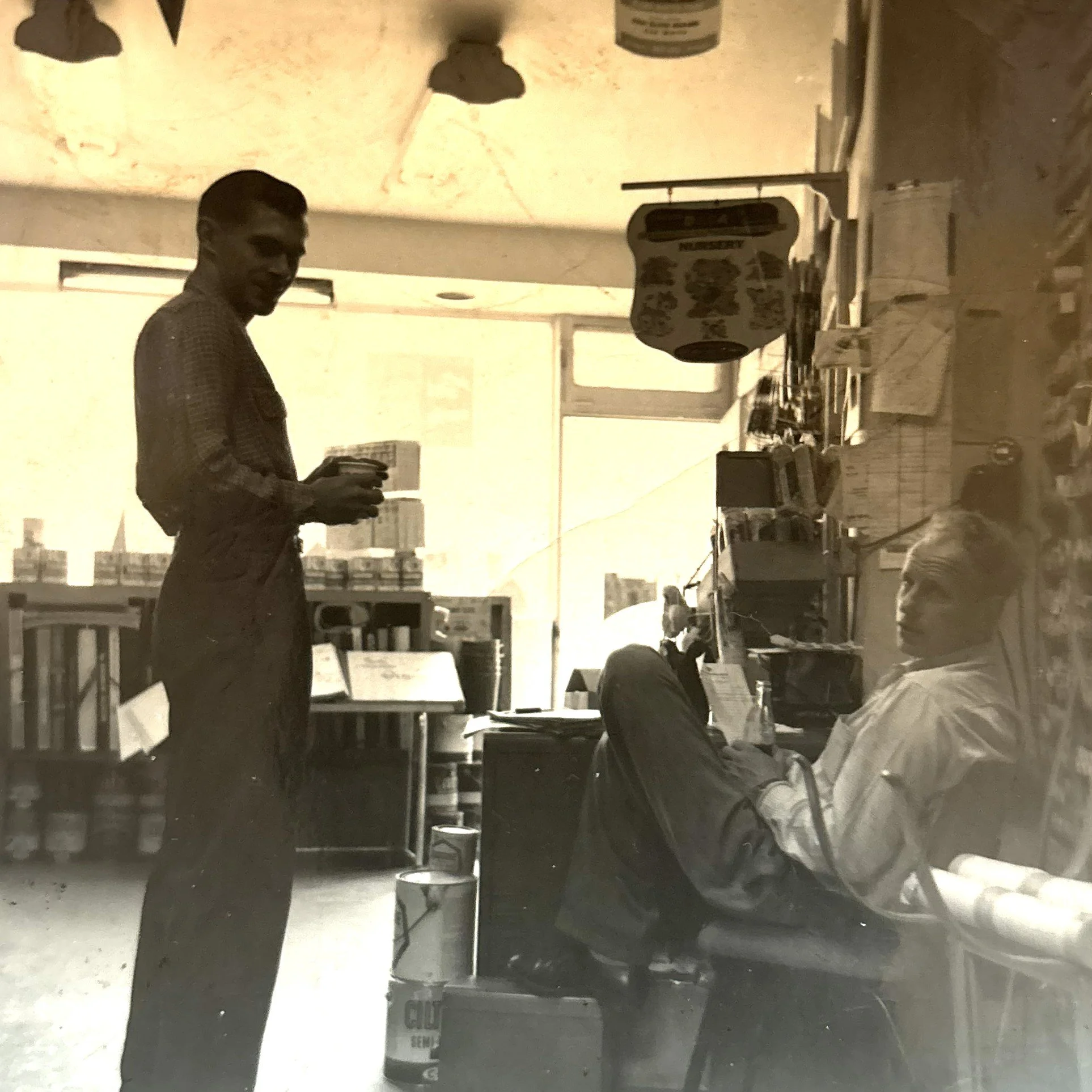 Black and white photo of two men inside a store. One standing man is holding an item and the other sitting man is engaged with a device. The store has shelves of products and a window in the background.