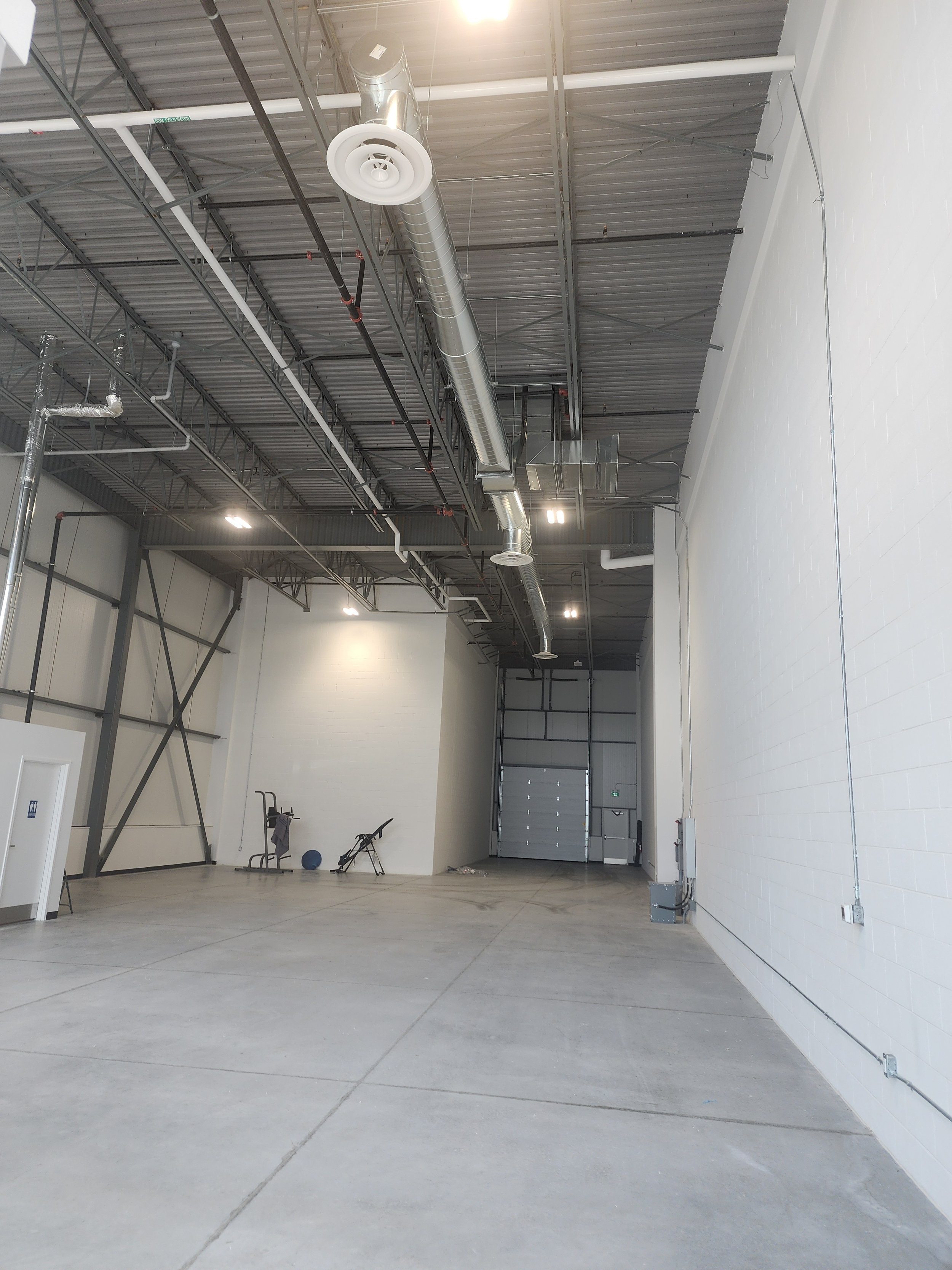Empty warehouse or industrial space with high ceiling, exposed ductwork, gray concrete floor, and minimal gym equipment including a treadmill, exercise ball, and a chair.