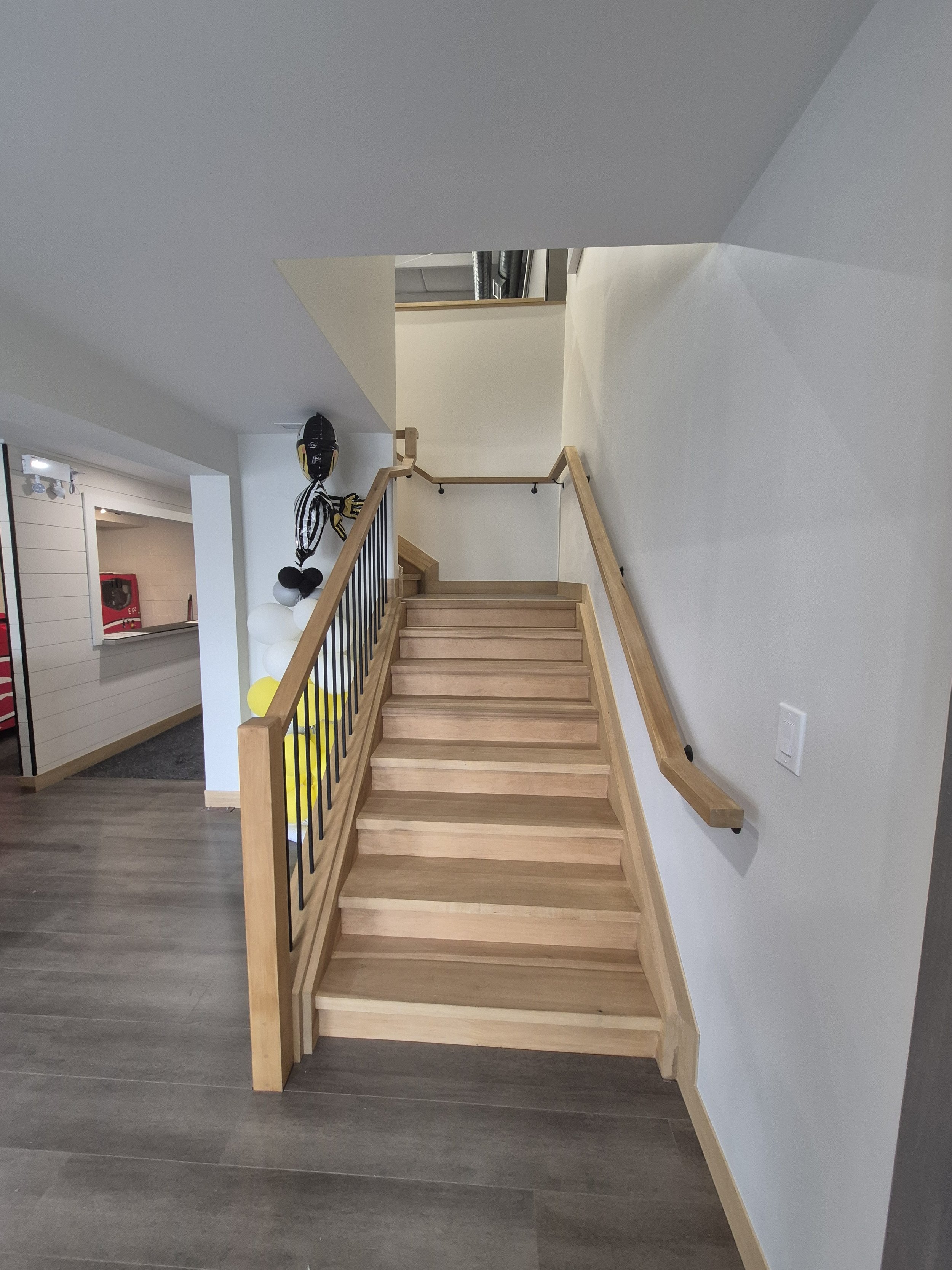 Wooden staircase with black iron railing and a beige handrail, leading upstairs in a modern interior space, with grey flooring and white walls. There are balloons and a large balloon figure shaped like a person in the background near the staircase.