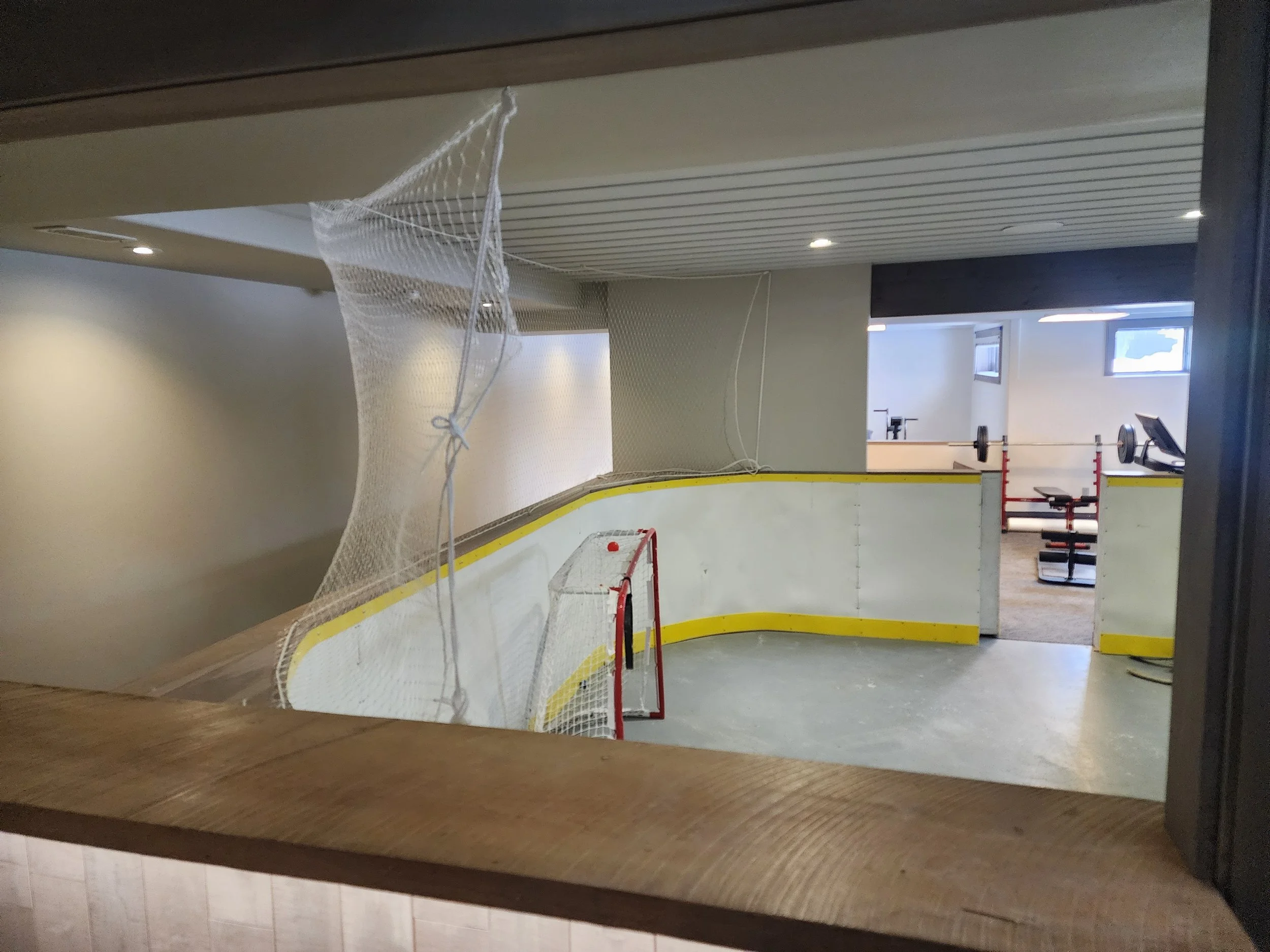 Indoor hockey rink with a small goal, barrier walls, and a net draped over part of the rink, overlooking a gym area with weight equipment.
