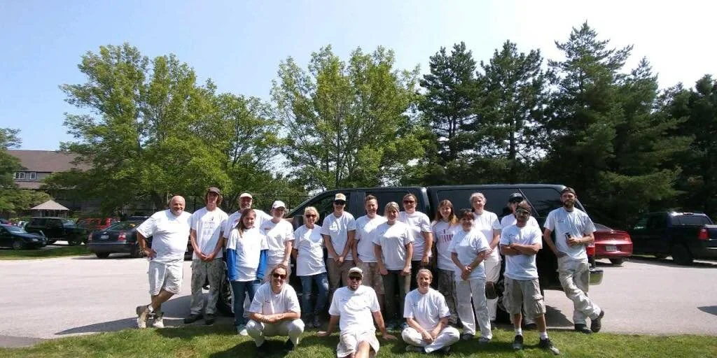 Group of people standing outdoors in a parking lot, with trees in the background, all wearing white clothing.