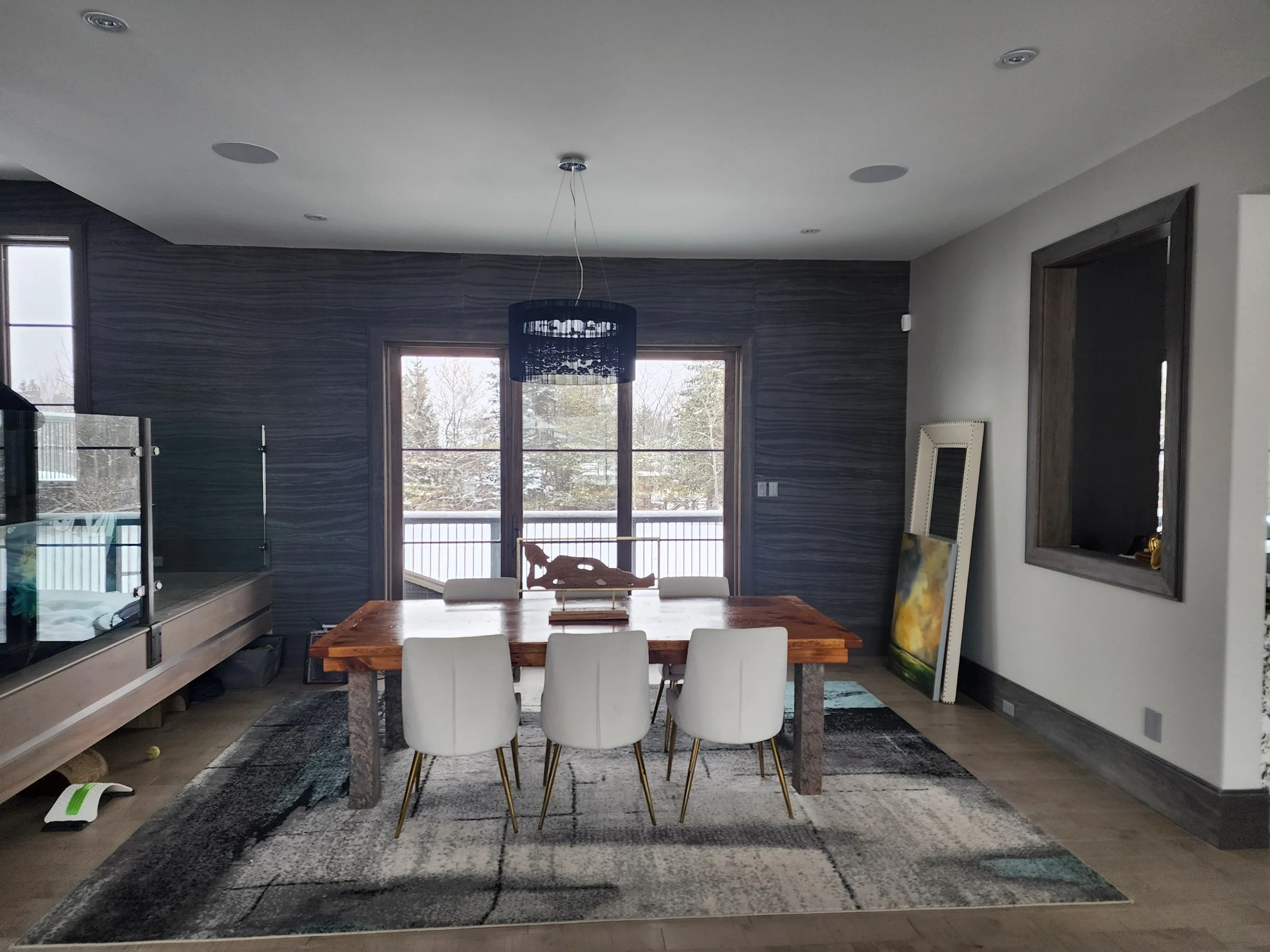Modern dining room with a wooden table, six white chairs, a dark blue hanging light fixture, large sliding glass doors showing snow outside, and artwork leaning against the wall.