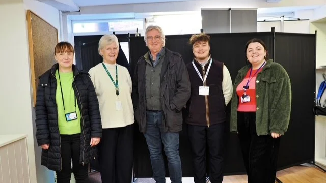 Group of five people standing side by side indoors, smiling for the camera, with black curtains behind them.