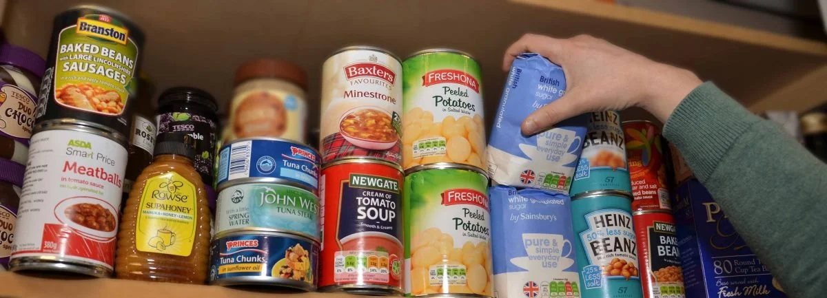 Shelf with canned foods and a hand reaching for a blue can of sugar.