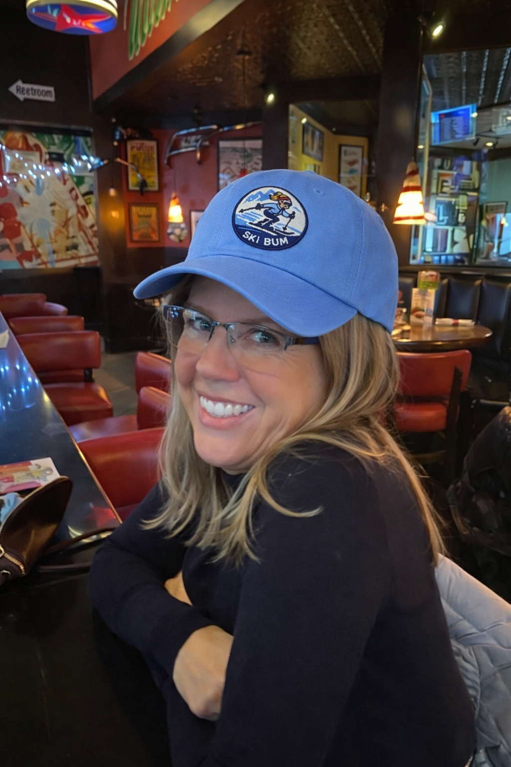 A woman smiling in a restaurant, wearing glasses and a blue baseball cap with a ski bum patch on it.