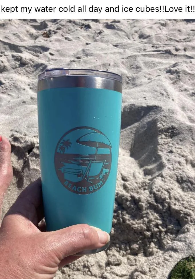Long weekend lifestyle-A person holding a tall blue tumbler with a beach-themed logo that includes an umbrella, a beach chair, and a palm tree, at the beach with sand in the background.