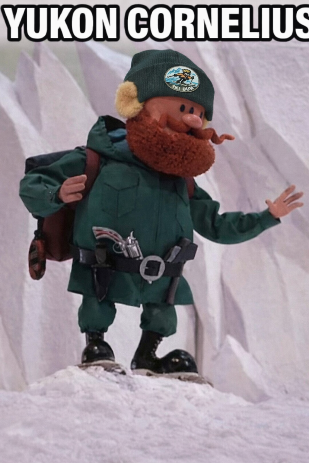 A plush toy with a cartoonish face, large nose, red beard, and wearing a beanie labeled Yukon Cornelius, dressed in winter clothing and standing on snow.