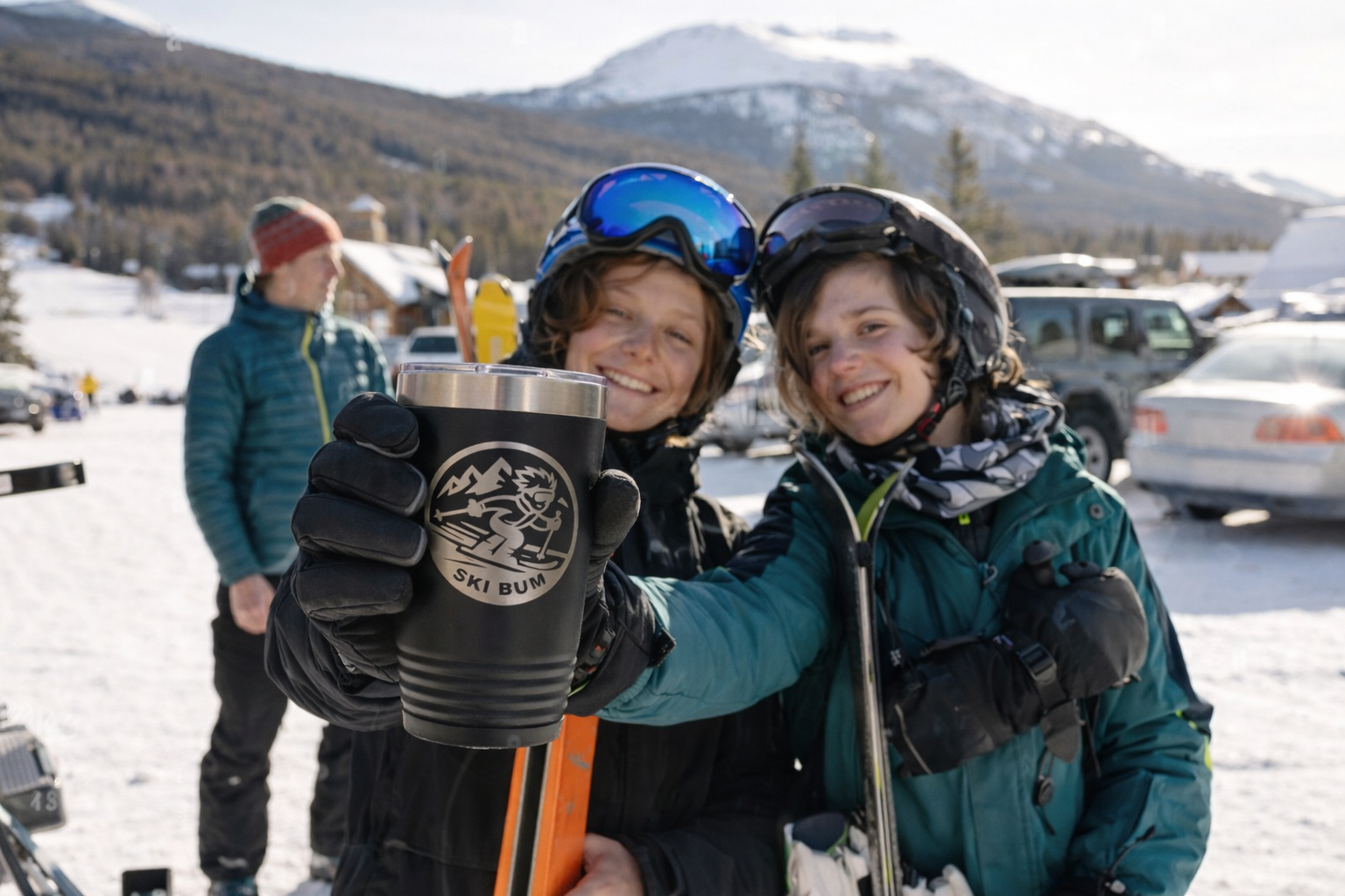 Two young male skiers smiling and cheerfully holding a cup with the logo 'Ski Bum' on it, outdoors in a snowy mountain landscape with cars and buildings in the background.