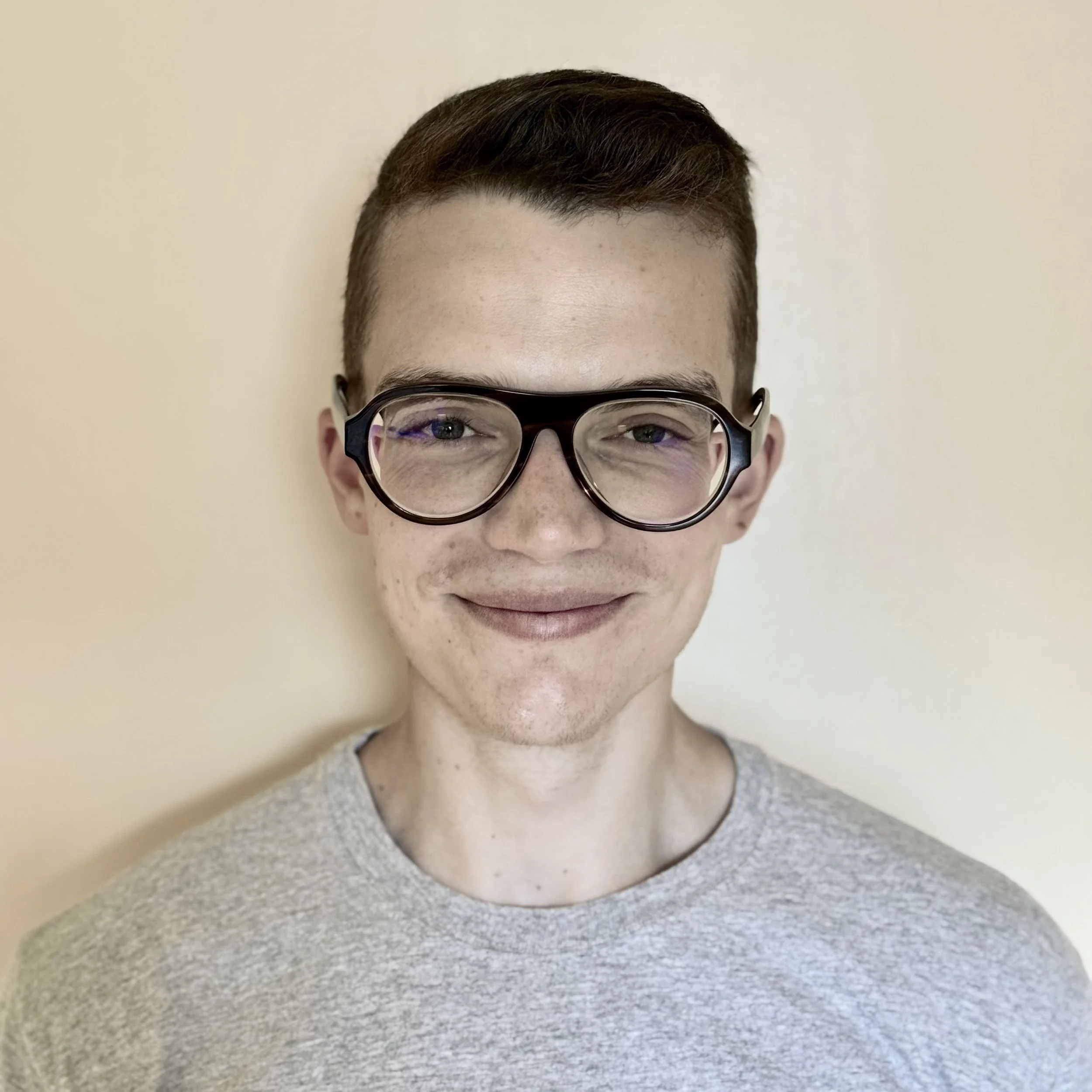 A young man with short brown hair, black glasses, and a light gray T-shirt smiling against a plain beige background.