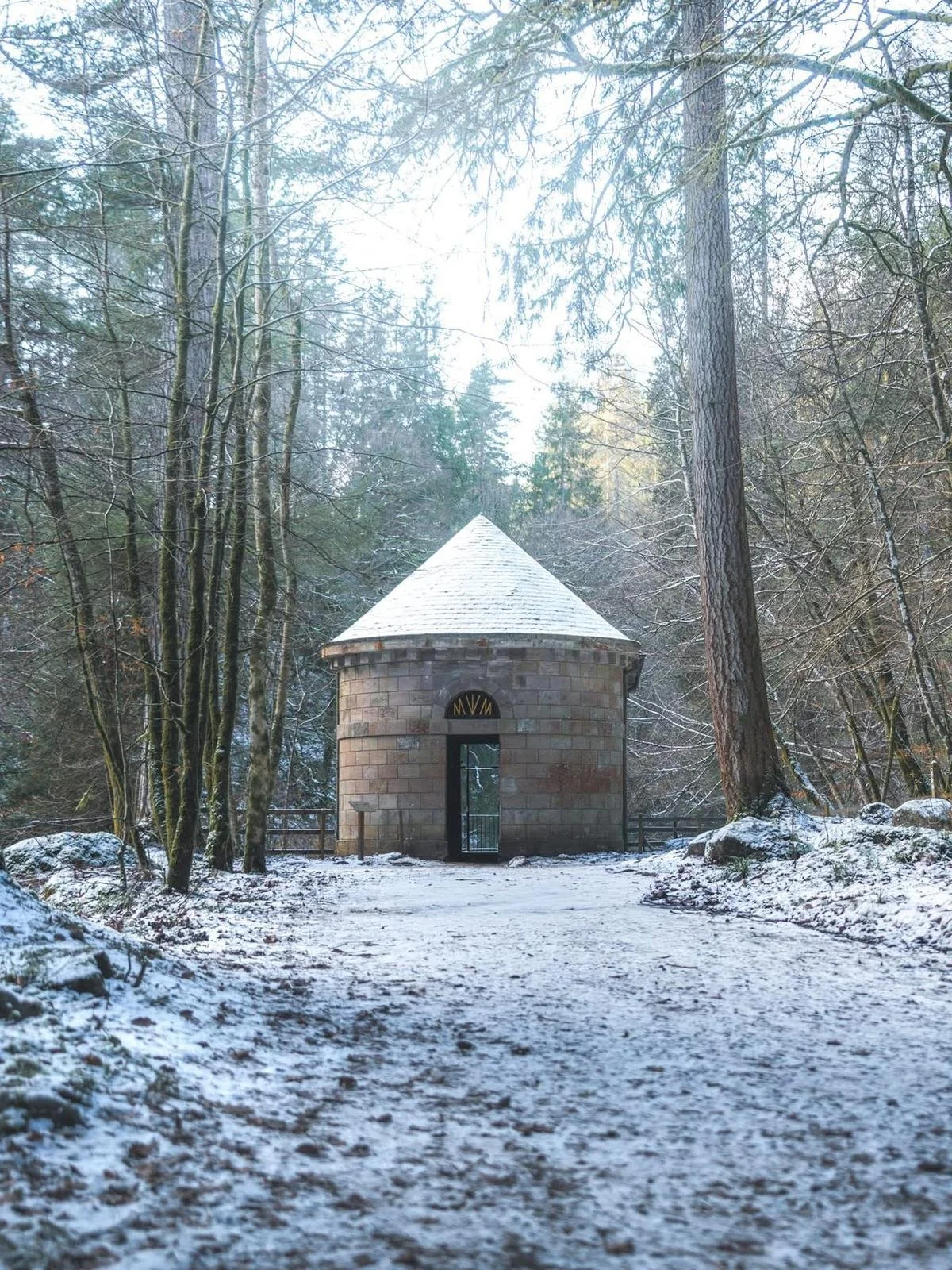 A small, round stone building with a cone-shaped roof in a snowy forest, with the entrance door visible and the letters 'NVM' above it.