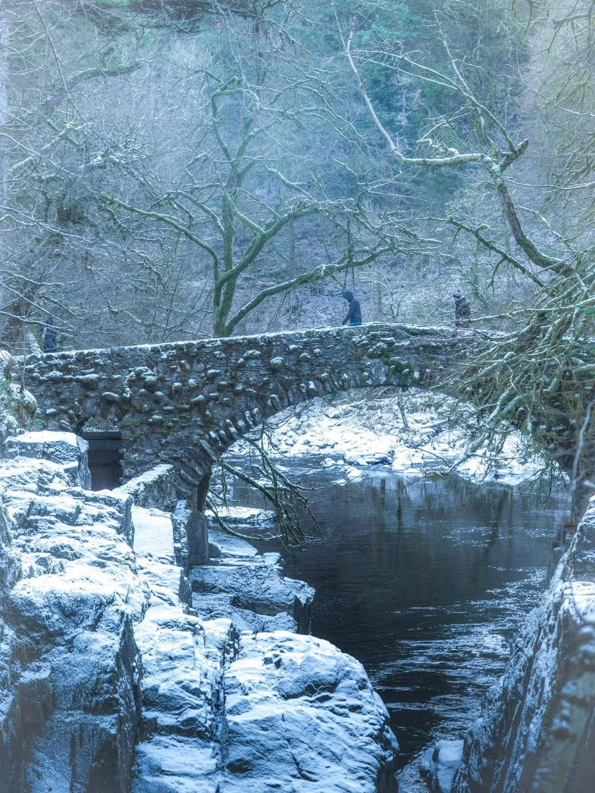 A snow-covered stone bridge over a river in a forest during winter. Two people are walking across the bridge, surrounded by snow and leafless trees.